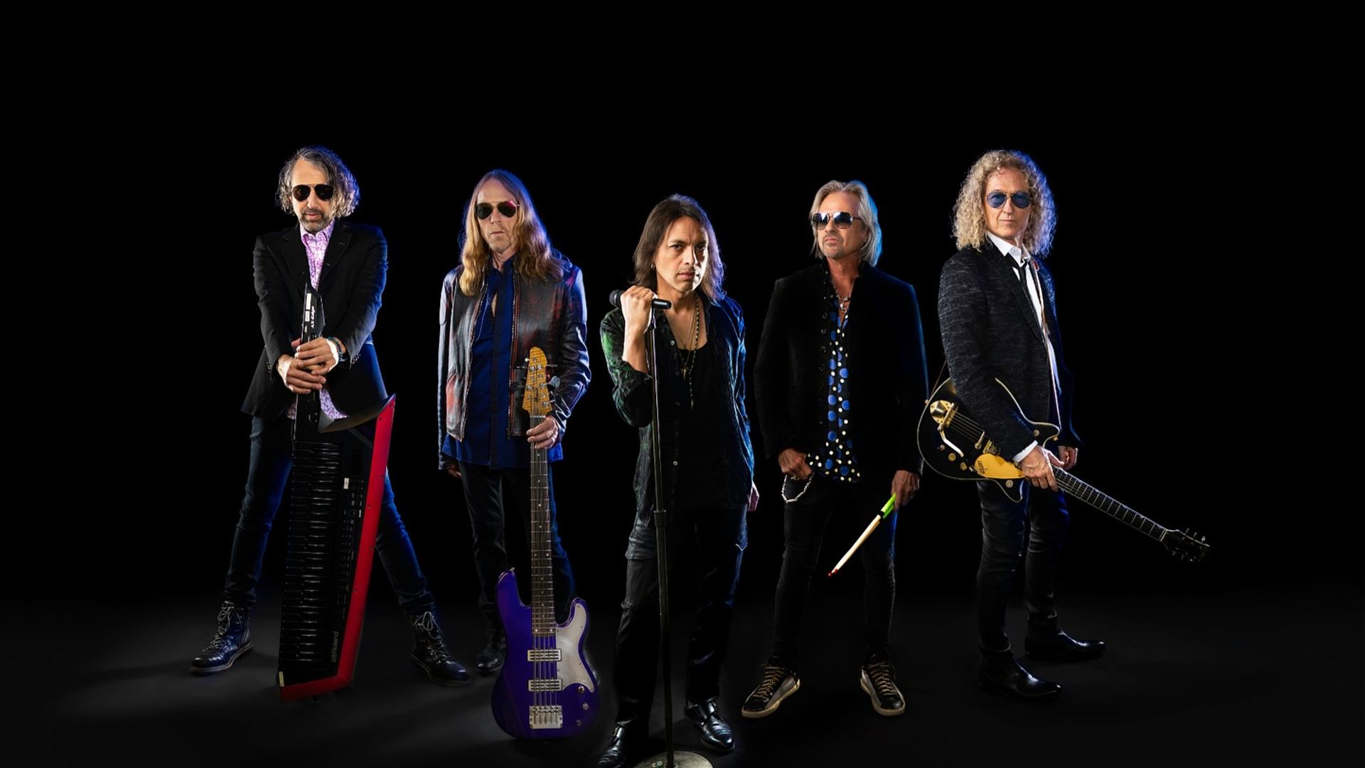 Rock band Foreigner posing with instruments against a black background.