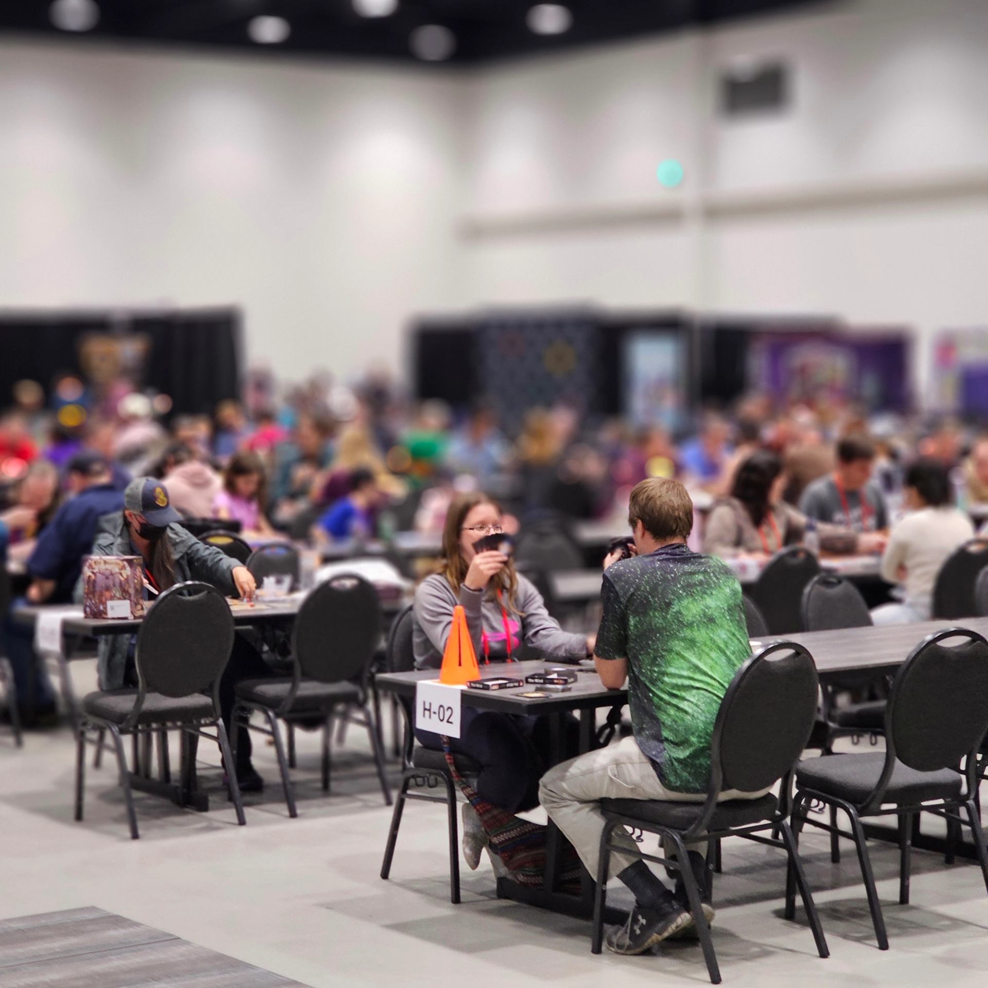 Crowded convention hall with people seated at tables playing card or board games.