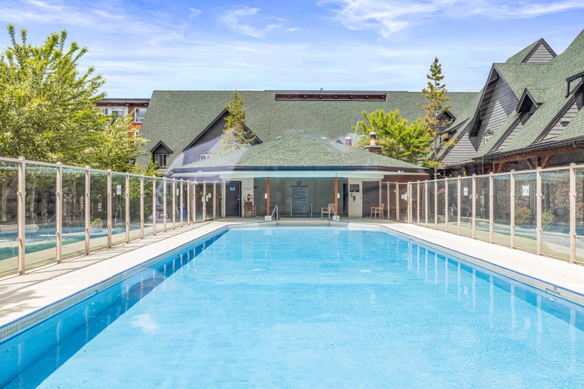 Outdoor swimming pool enclosed by glass fencing beside lodge-style buildings and trees.
