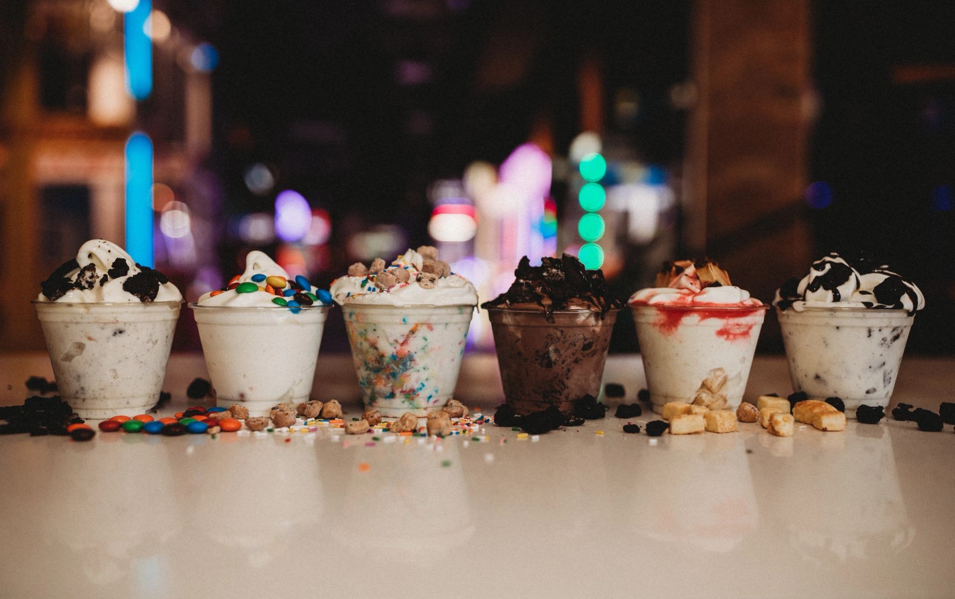 Ice cream sundaes in clear cups with toppings on a table, blurred background lights.
