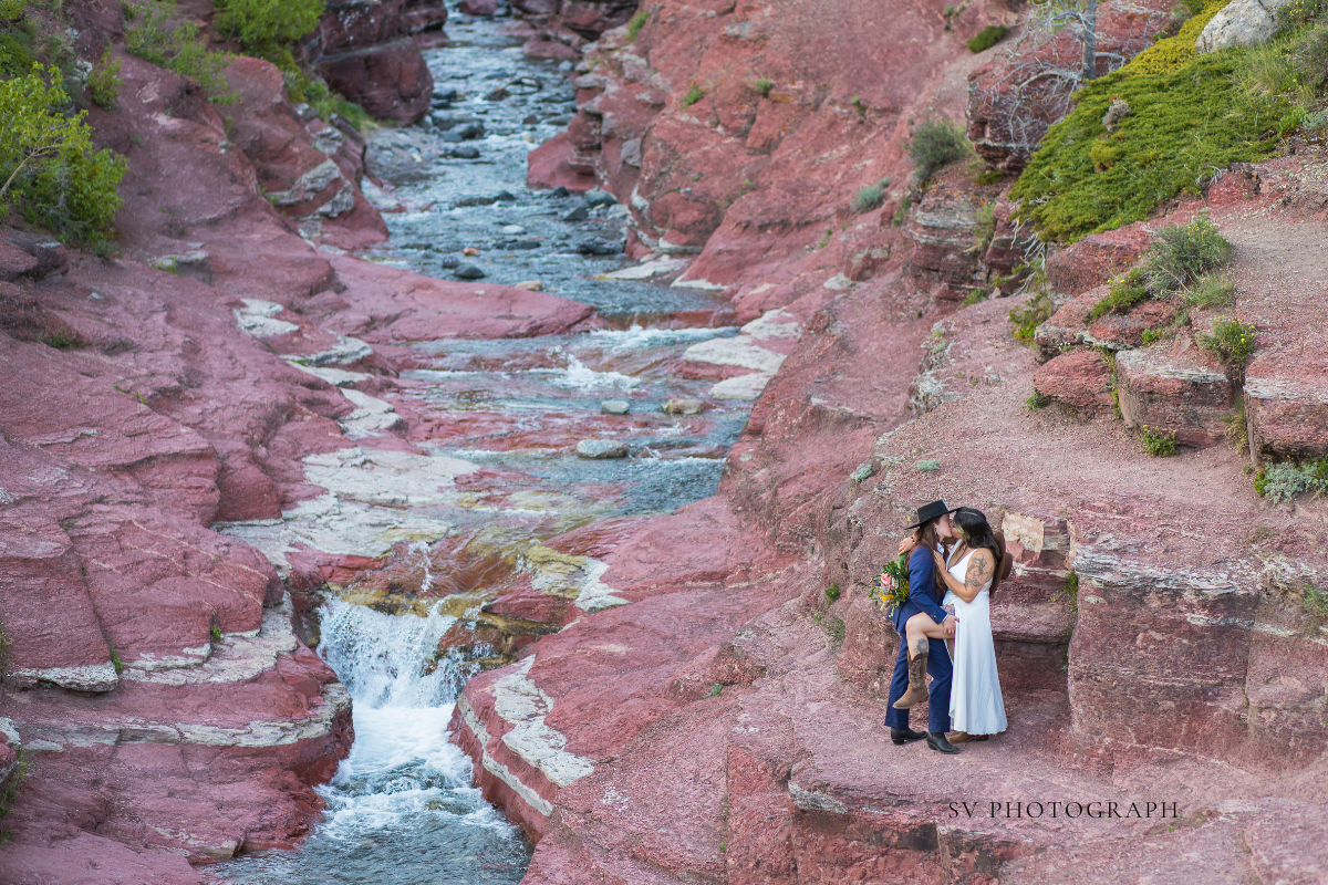 A couple embraces on a red rock canyon ledge beside a flowing creek and small waterfall.