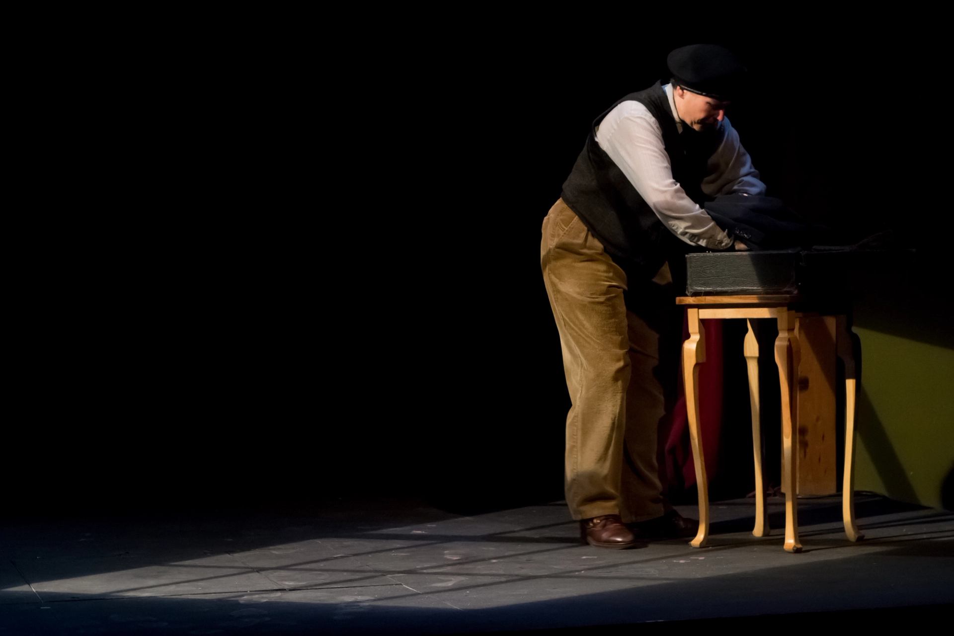 Stage performer in vintage clothing leans over suitcase on small wooden table.