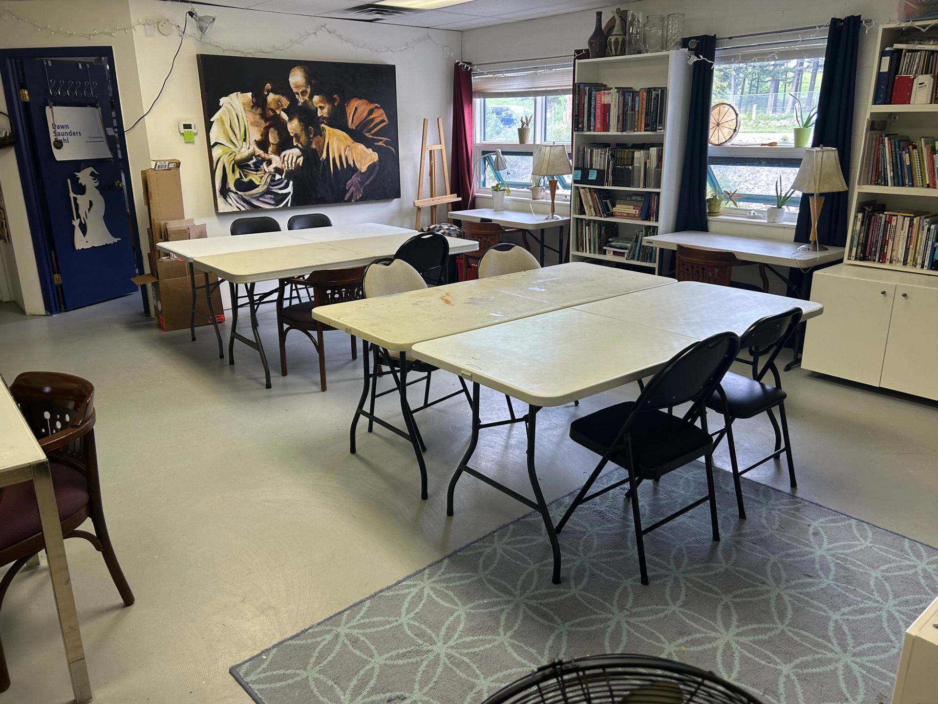 Bright studio with tables, bookshelves, and a large wall painting.