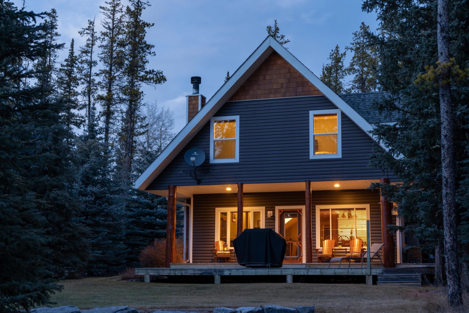 A-frame lodge with lit windows, front deck chairs, and tall pine trees nearby.