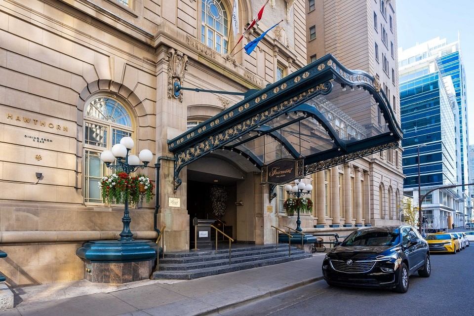 Elegant hotel entrance with glass canopy, ornate details, and flags. Steps lead to a grand doorway. A black car is parked in front on a city street.