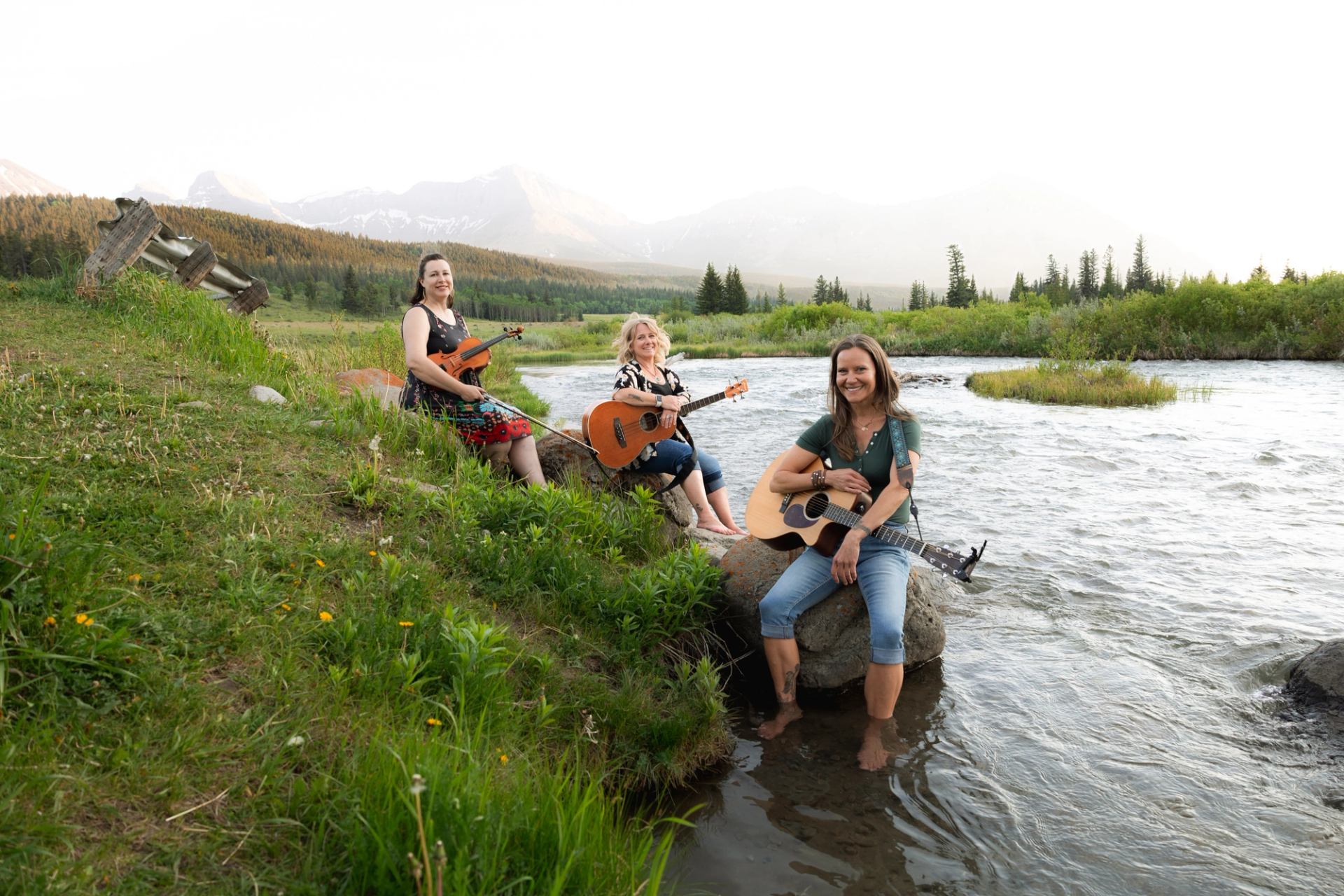 Three women with a fiddle and two guitars sit by a river in a mountain landscape.