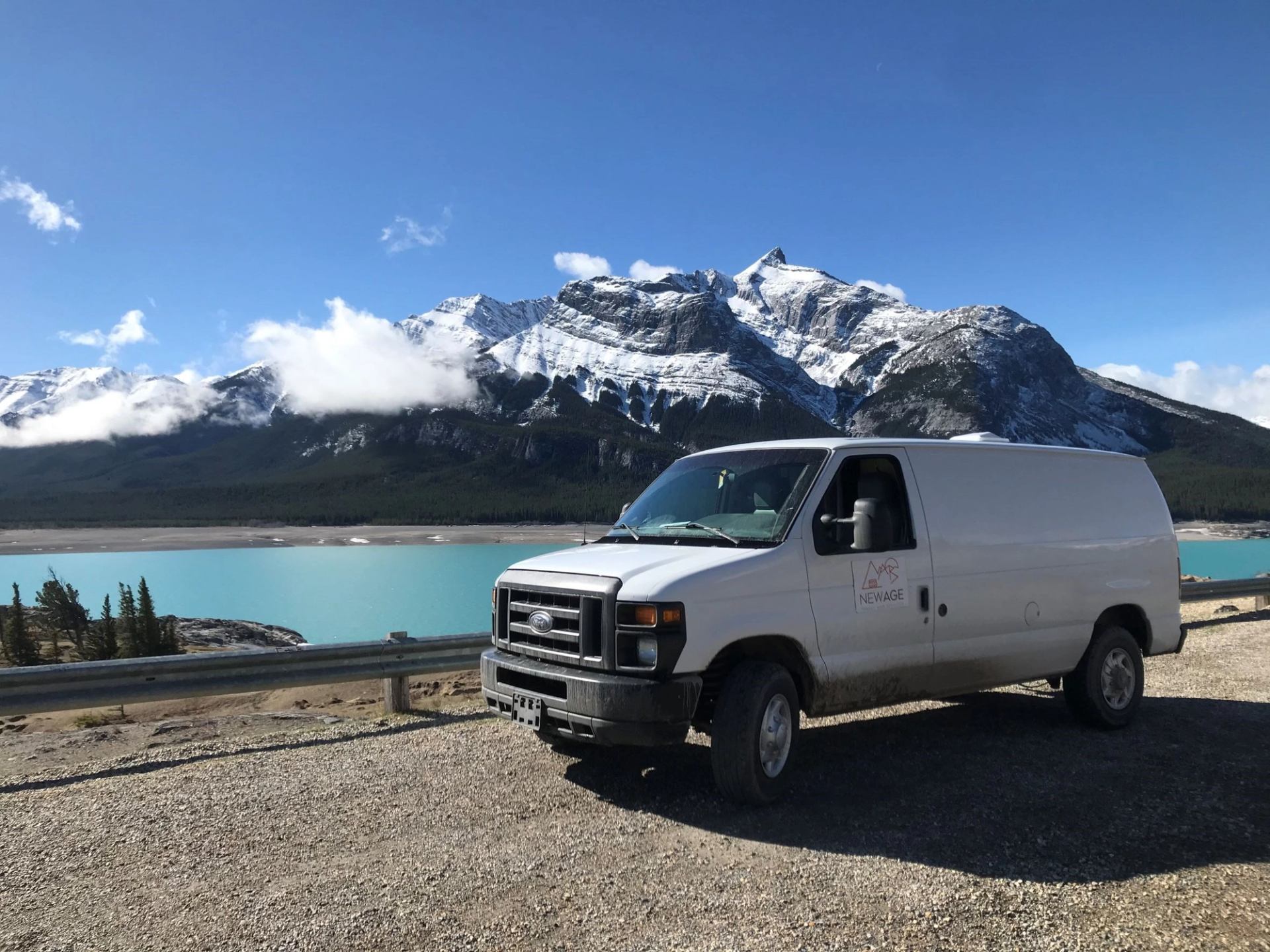 White van parked by turquoise lake with mountain backdrop.