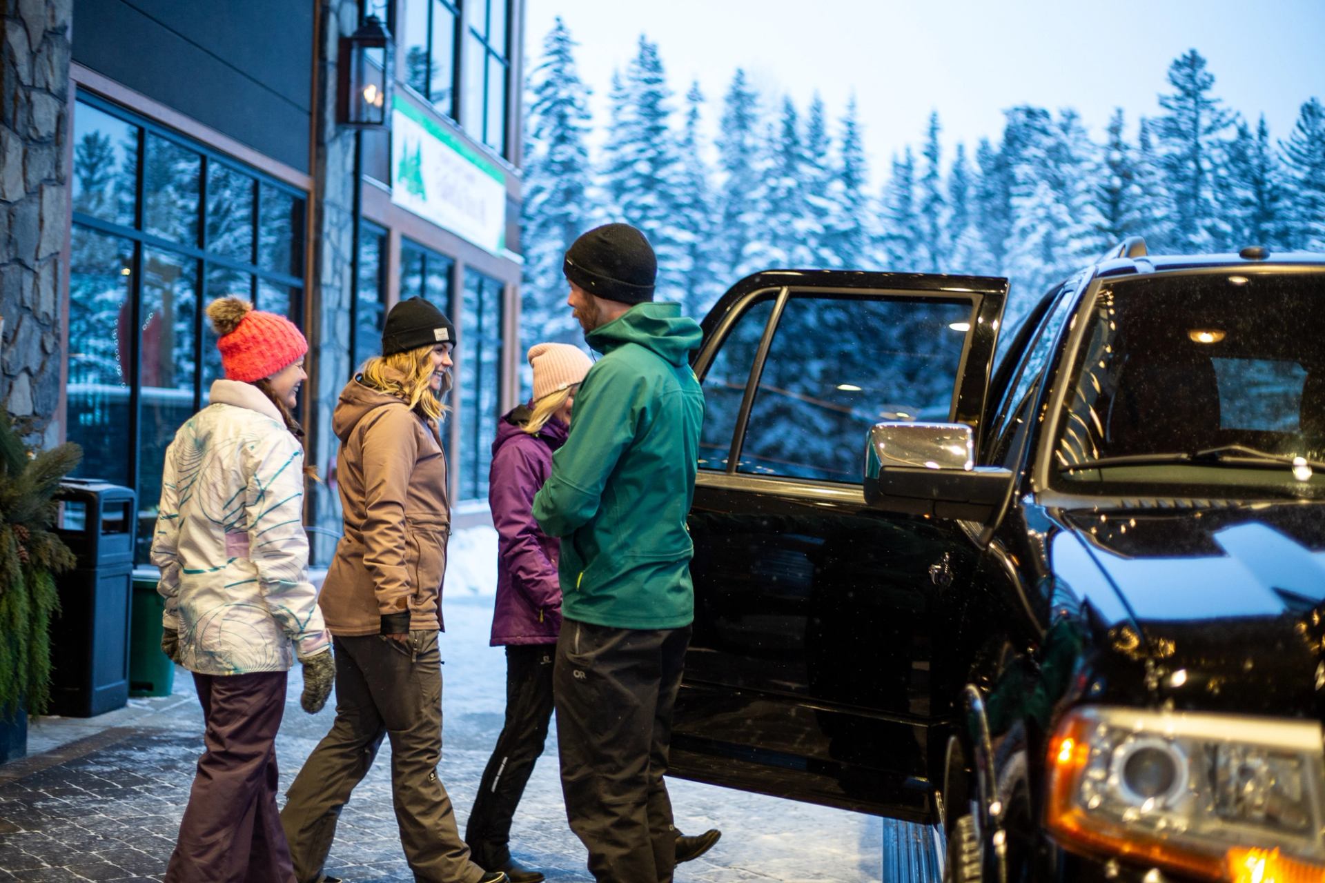 Group preparing to enter a vehicle outside a lodge surrounded by snowy trees.