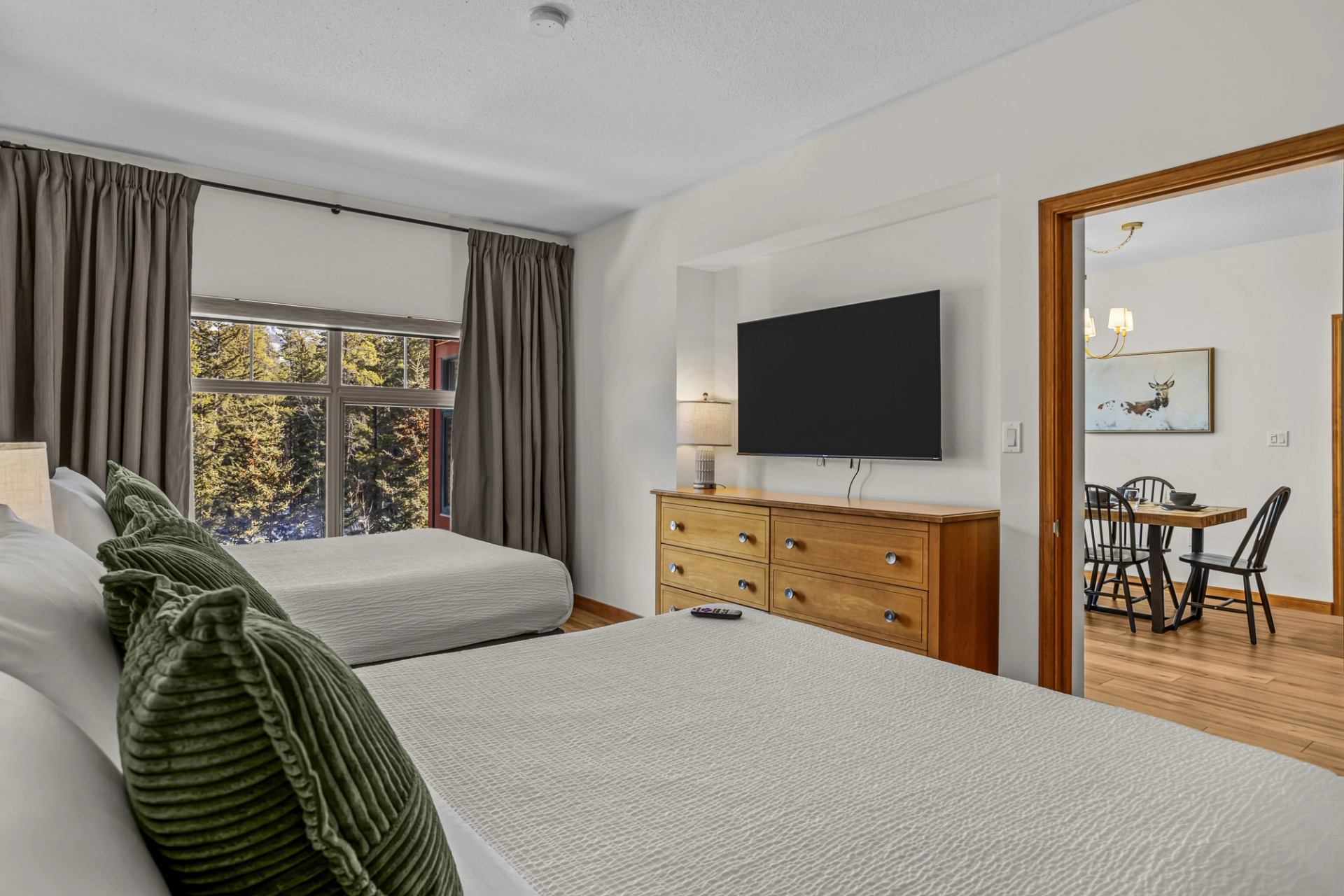 A bright bedroom features two neatly made beds, a large TV on a dresser, and a window overlooking tall evergreen trees.