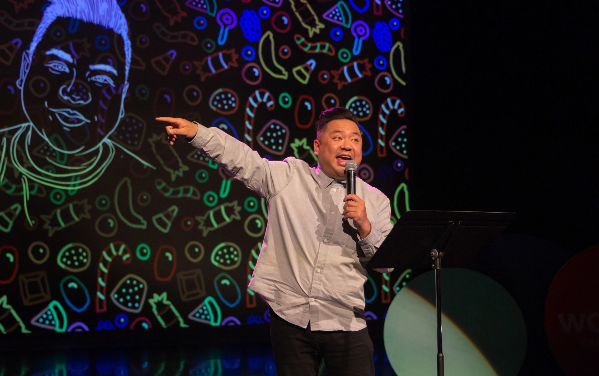 A speaker stands at a microphone on a theatre stage, pointing toward the audience while colourful illustrated artwork glows on a large screen behind them.