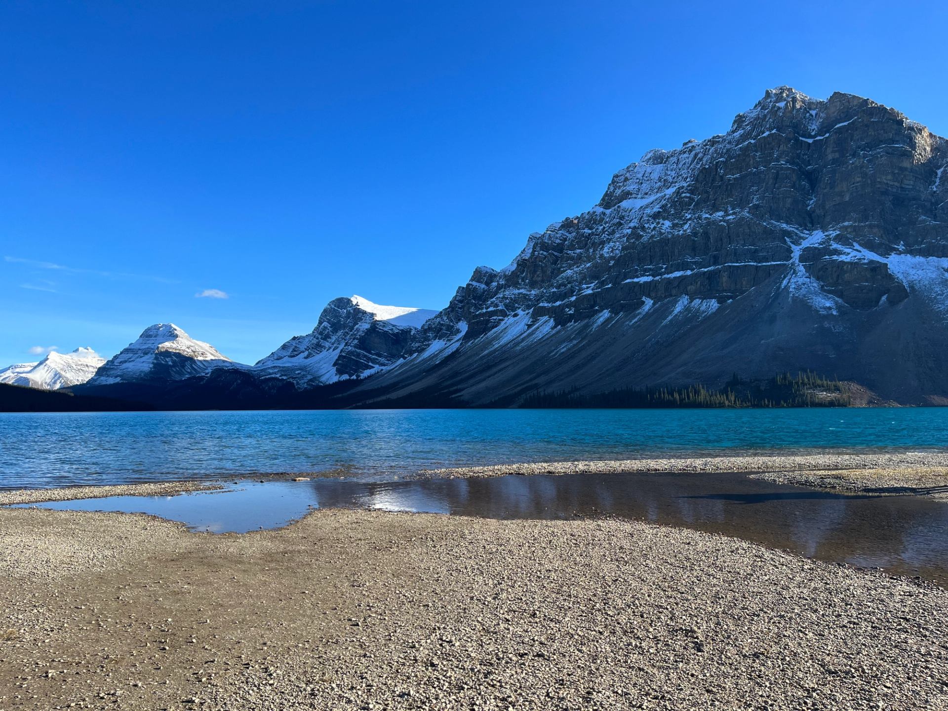 Rocky shoreline with turquoise lake and snow-dusted mountains under clear blue sky.