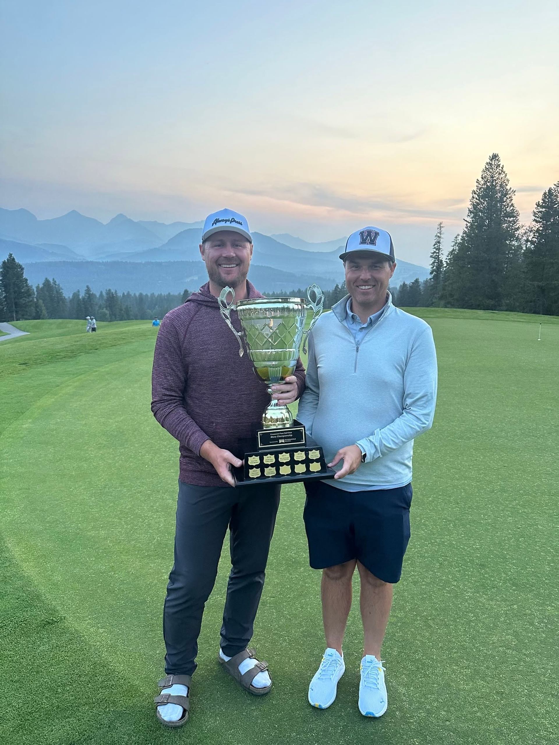 Two golfers stand on a green holding a large trophy, mountains in background.