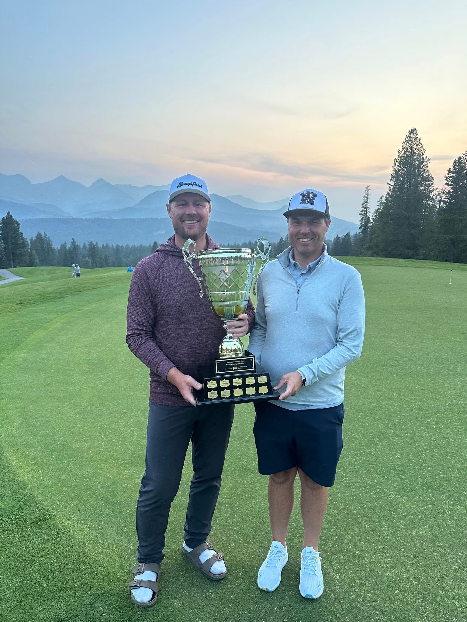 Two golfers stand on a green holding a large trophy, mountains in background.