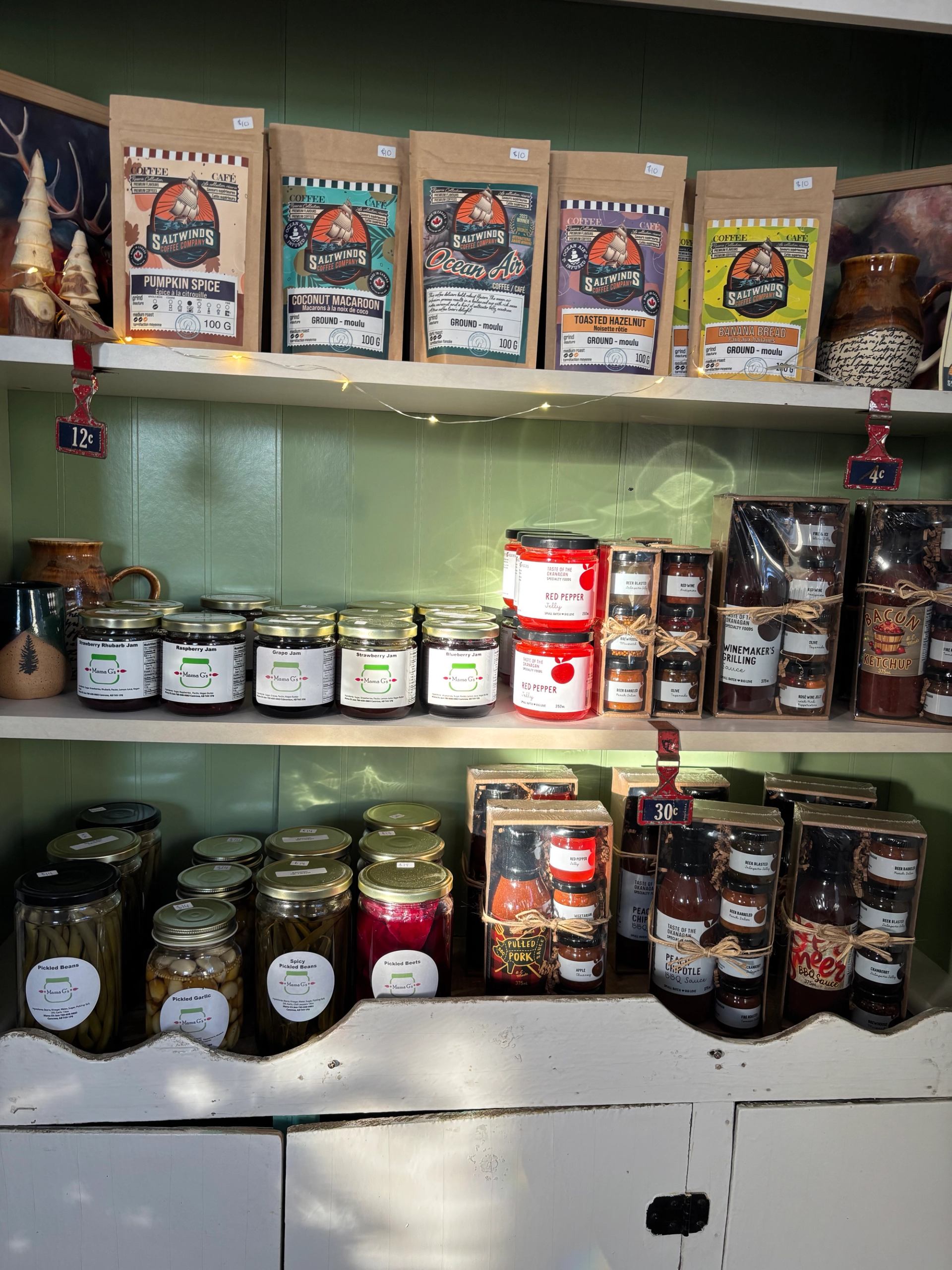 A rustic shelf display featuring various artisanal food products, including Saltwinds Coffee bags, jars of jams and pickled goods, and gift sets of sauces and spices.