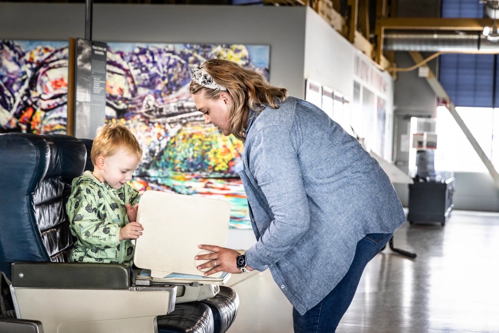 A woman shows a light-colored object to a young child seated in an airplane seat, with colorful abstract art on walls in the background.