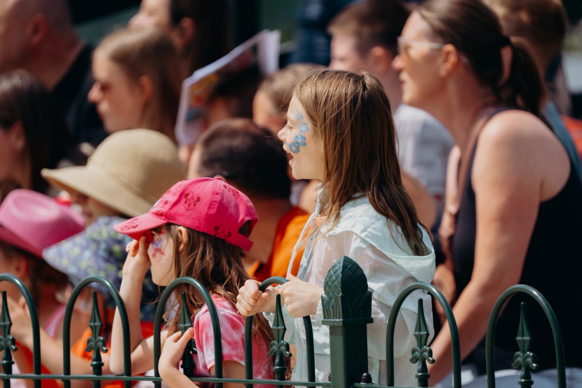 Children with face paint stand by a fence, watching an event in a sunny crowd.