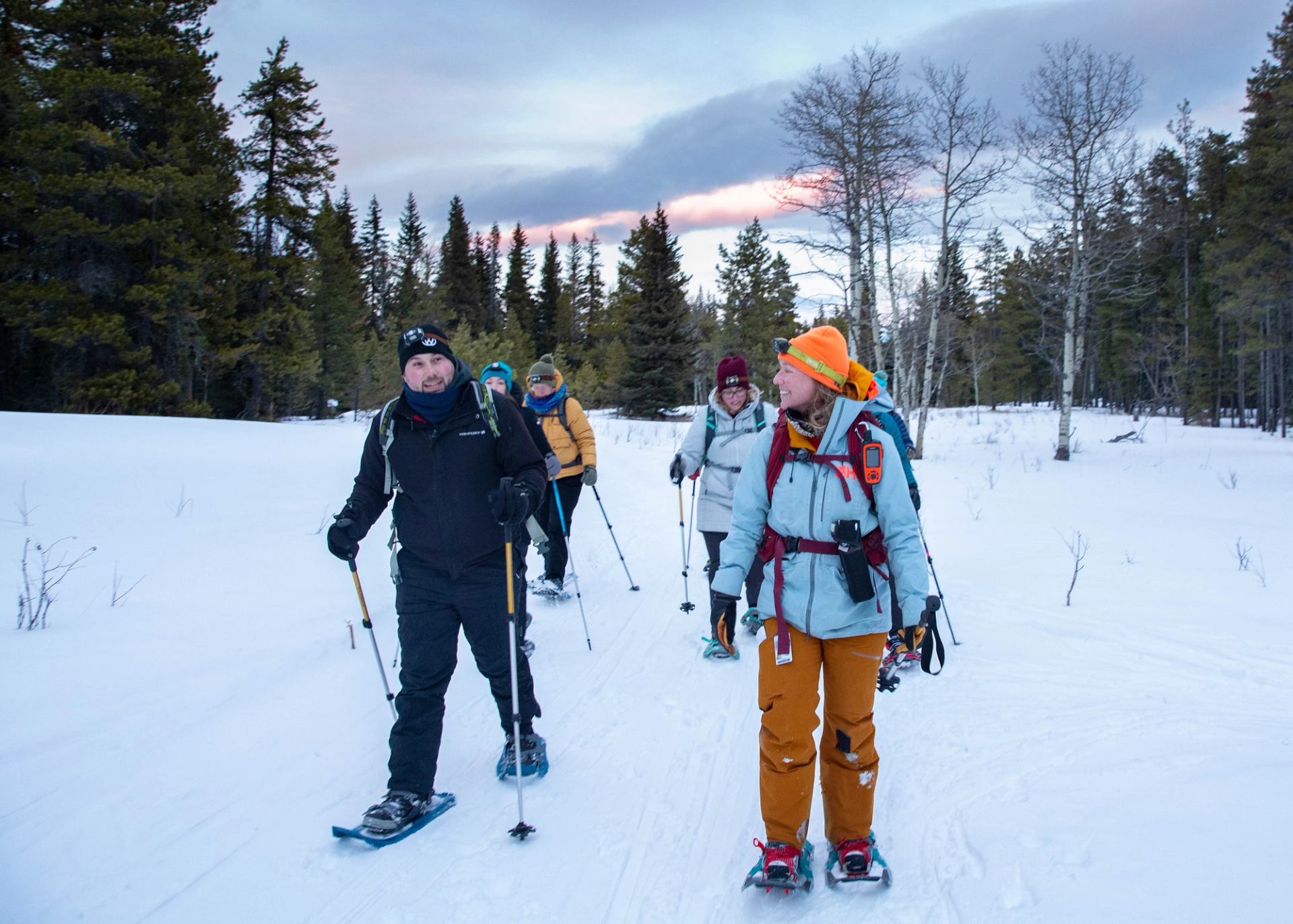 Group snowshoeing on a snowy trail at dusk with trees and a colorful sunset sky.