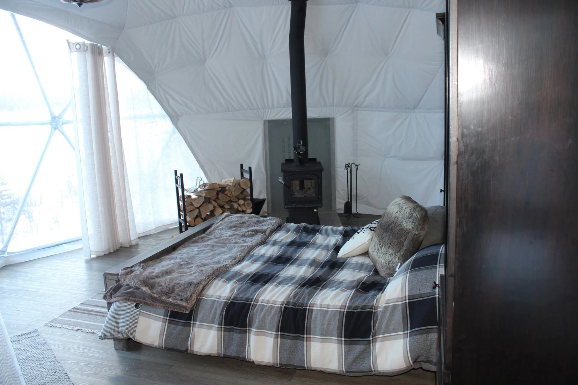 Cozy dome interior with plaid bed, wood stove, stacked firewood and sheer curtains.