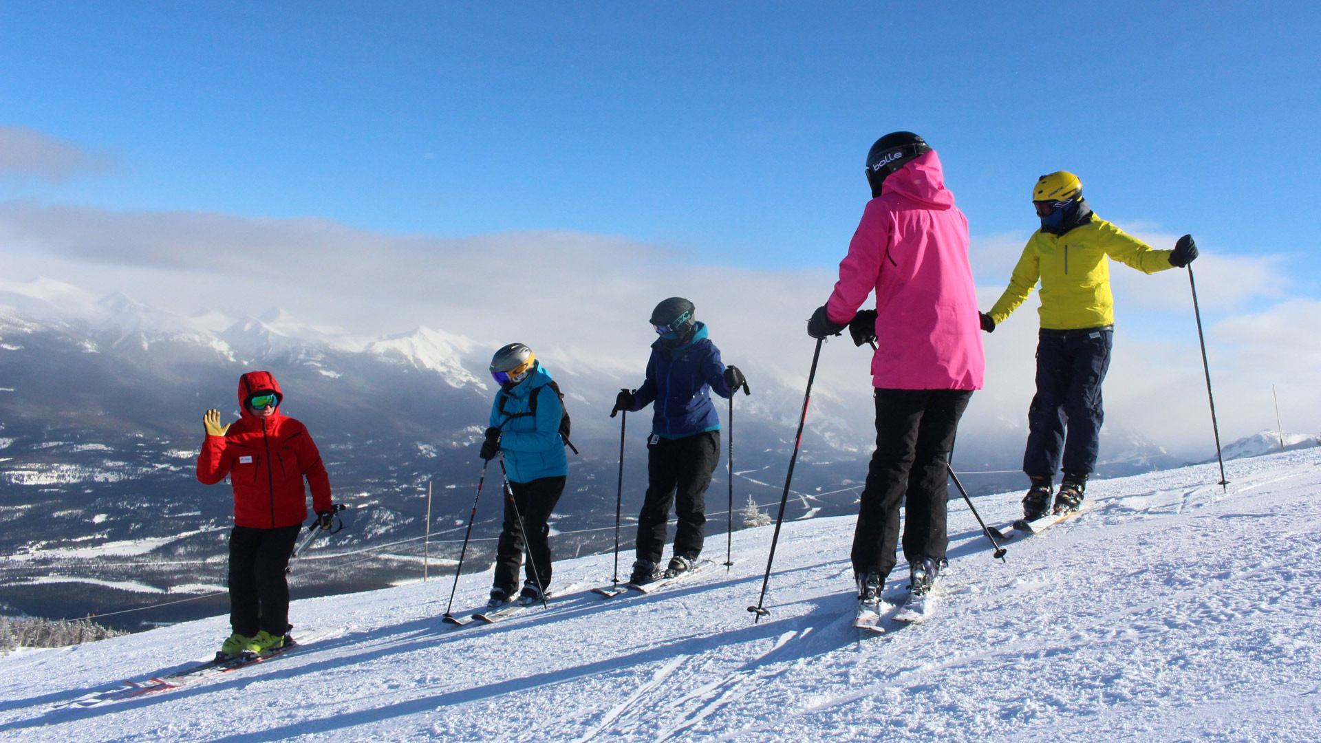 Group of skiers standing on a snowy slope with ski poles and mountains in the background.