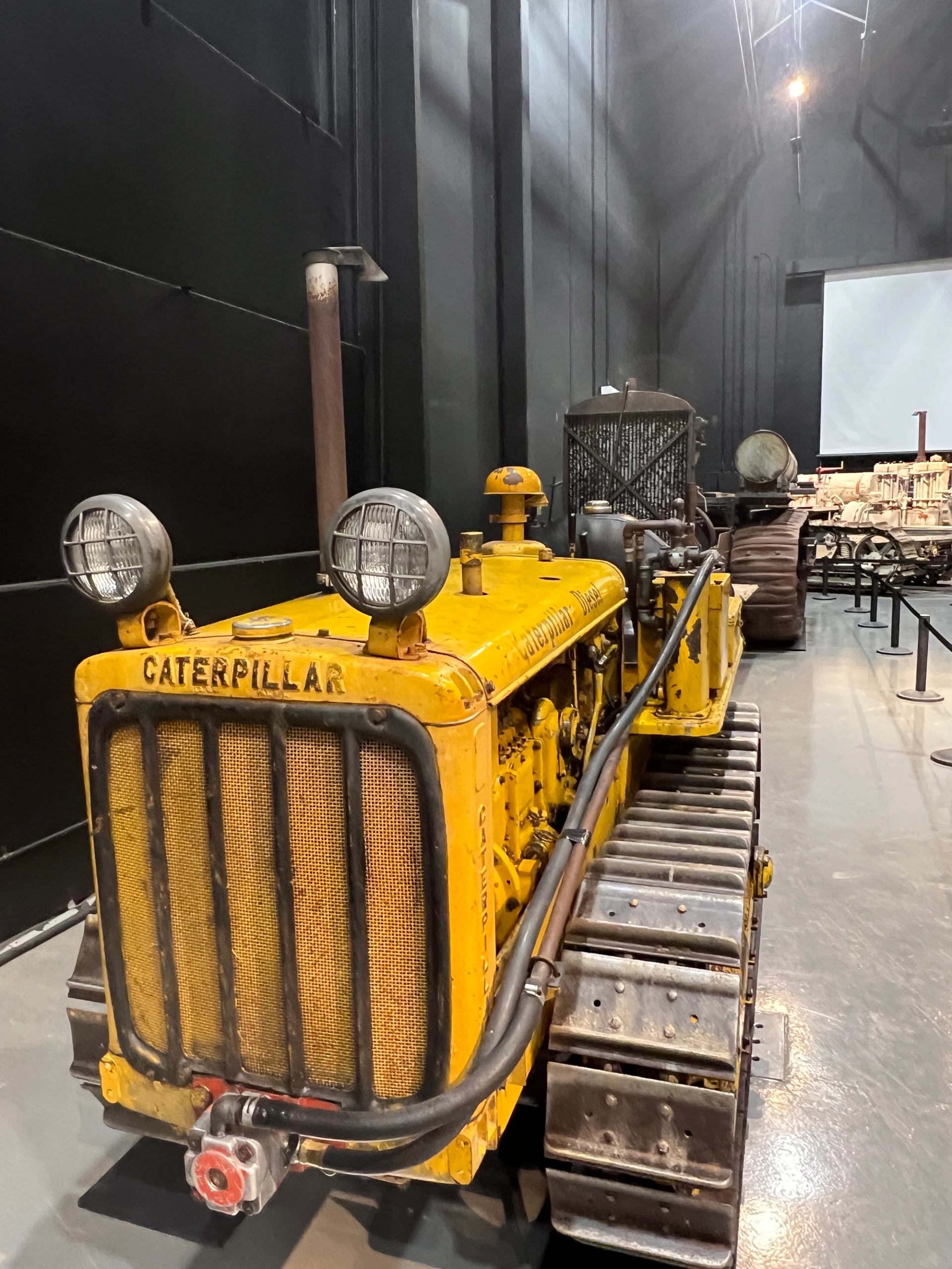 Yellow Caterpillar crawler tractor with headlights, shown in a museum exhibit hall.