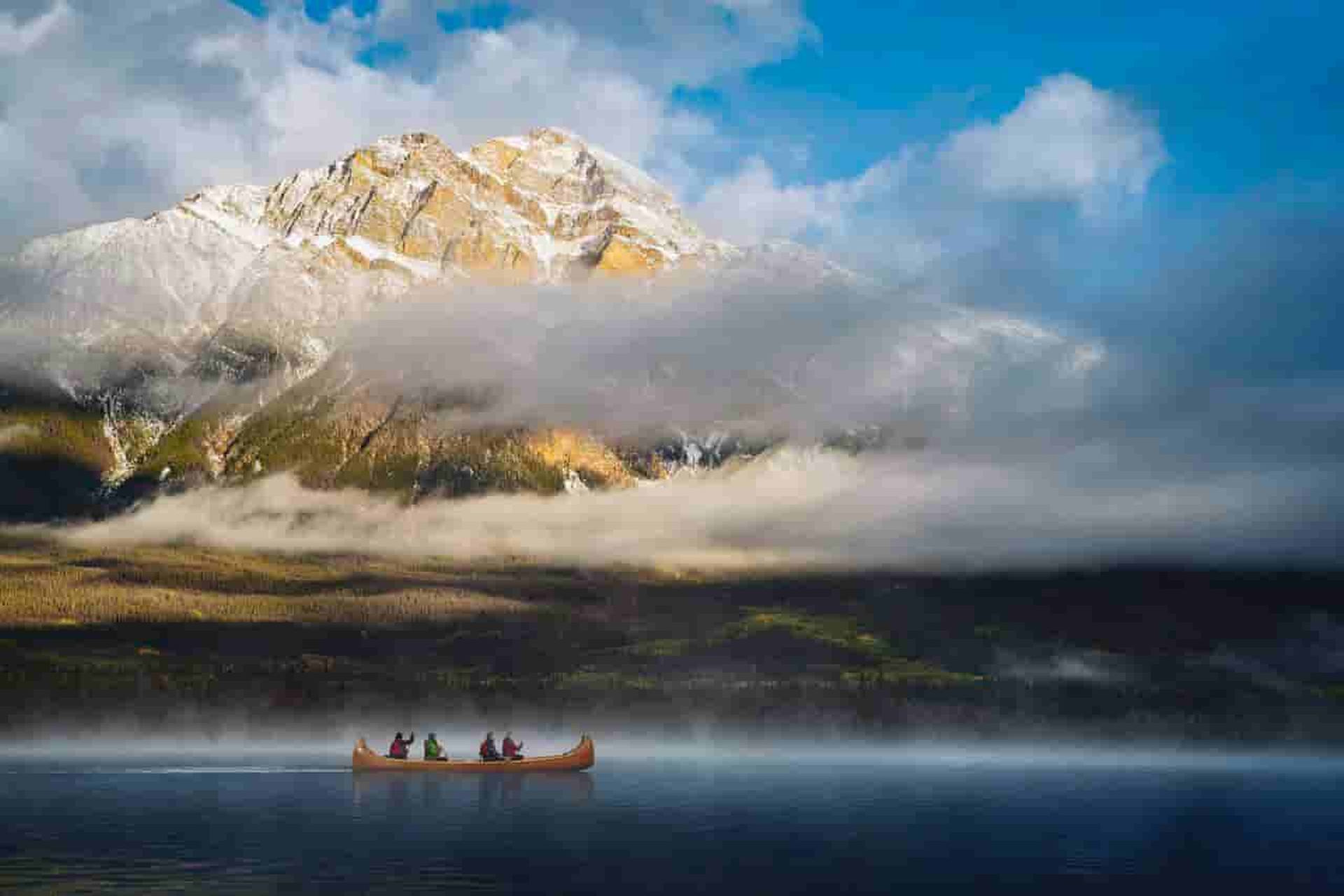 Canoe with paddlers on calm lake below misty, snow-dusted mountain and cloudy sky.