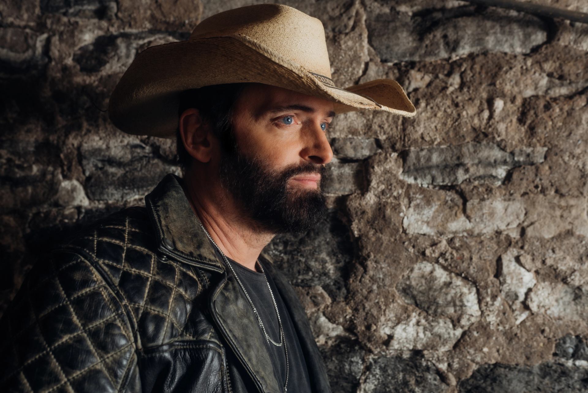 Bearded person in a cowboy hat and leather jacket stands beside a stone wall.