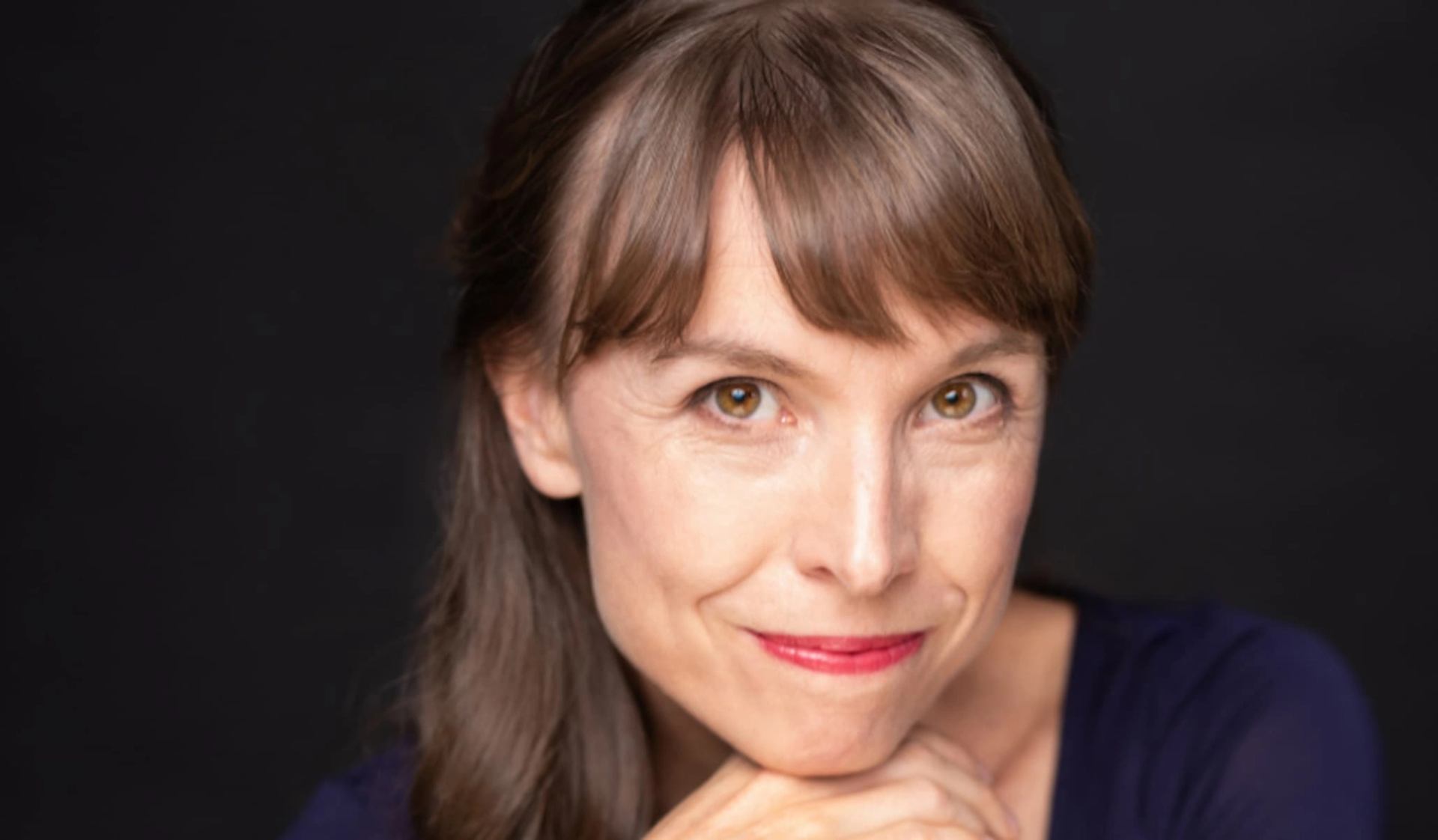 Person with brown hair and red lipstick resting chin on hands against dark background.