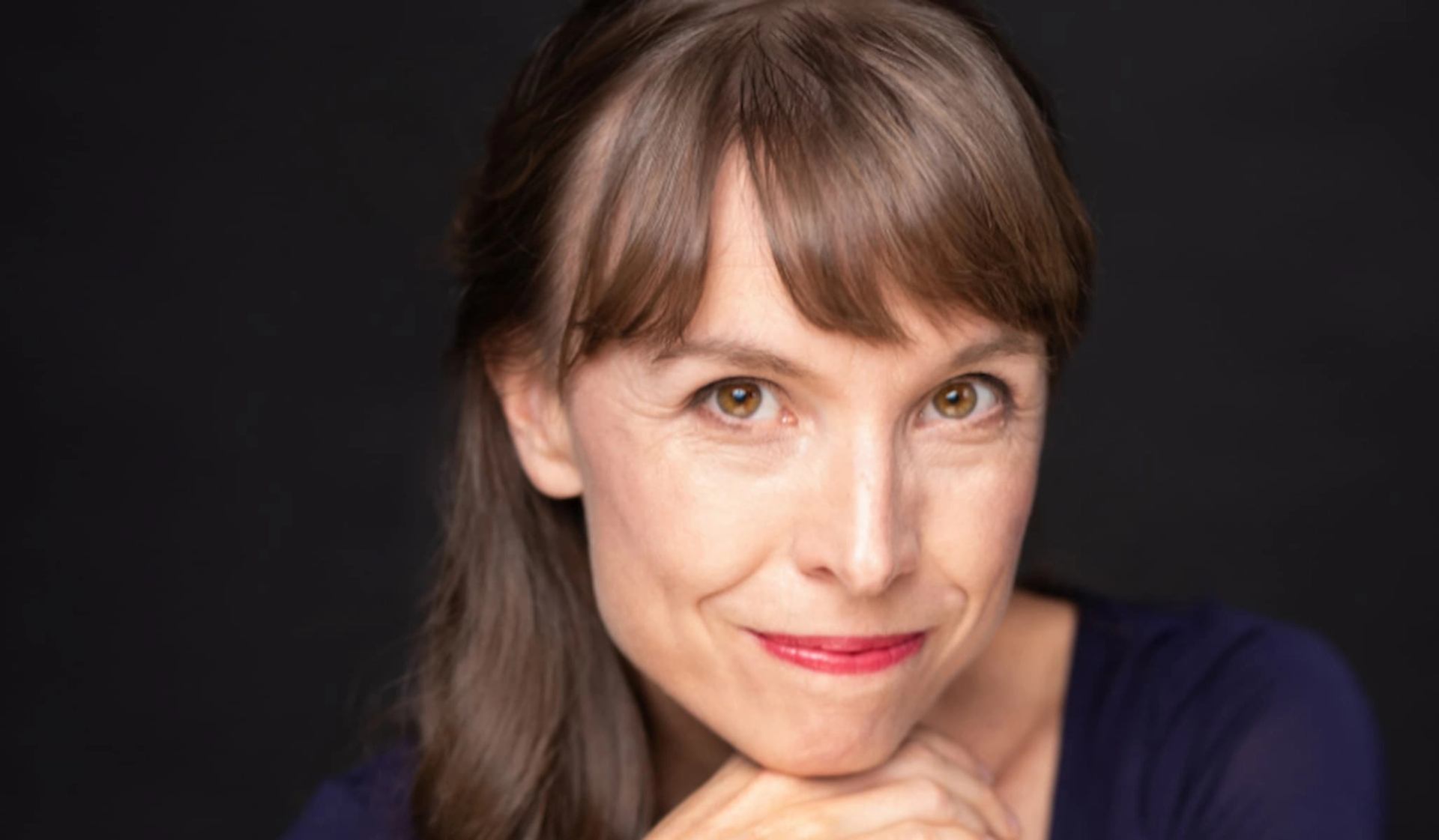 Person with brown hair and red lipstick resting chin on hands against dark background.