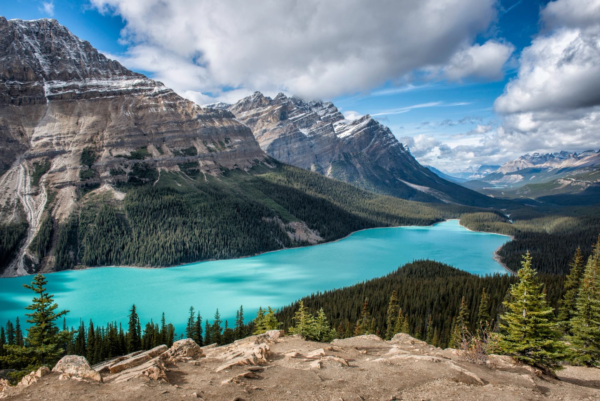 Turquoise mountain lake curves below forested slopes and rocky peaks under scattered clouds.