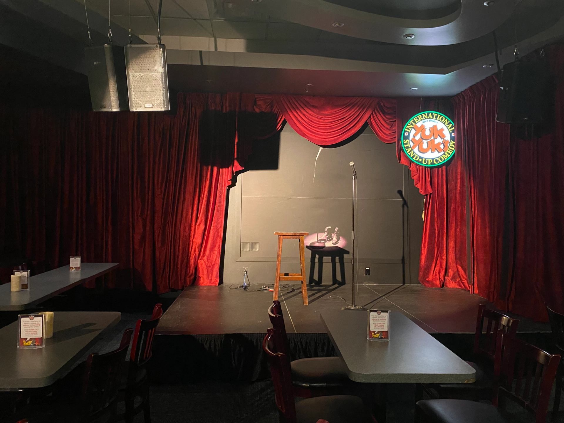 Small comedy stage with red curtains, mic stand, and audience tables.