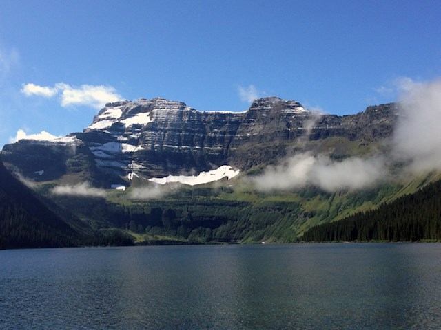 Calm lake with snow-capped mountains and lush greenery under a clear blue sky.