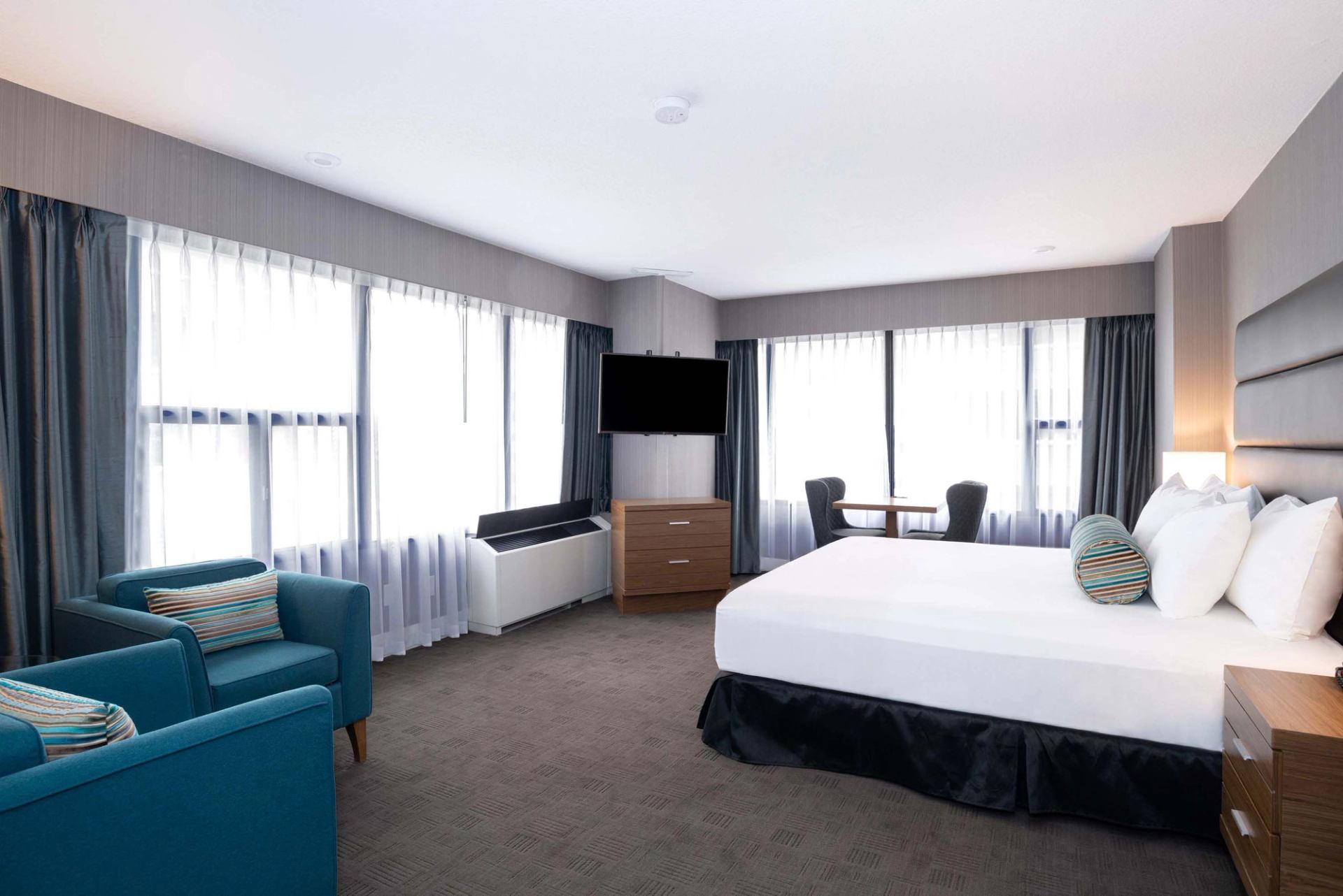Hotel room with king bed, teal armchairs, wall-mounted TV and large curtained windows.
