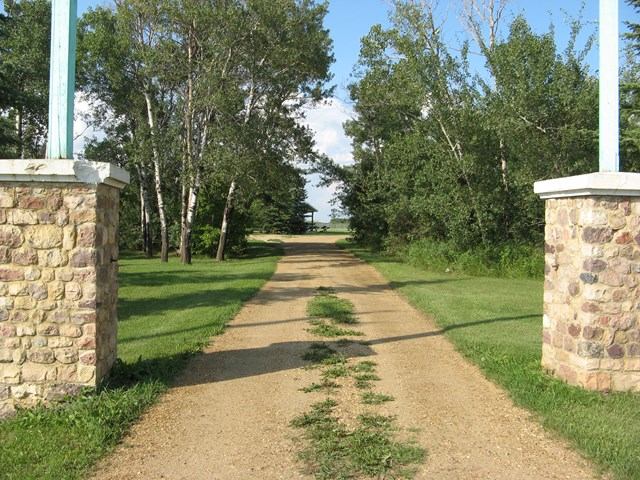 Stone-pillared entrance with dirt road leading into a tree-lined campground.