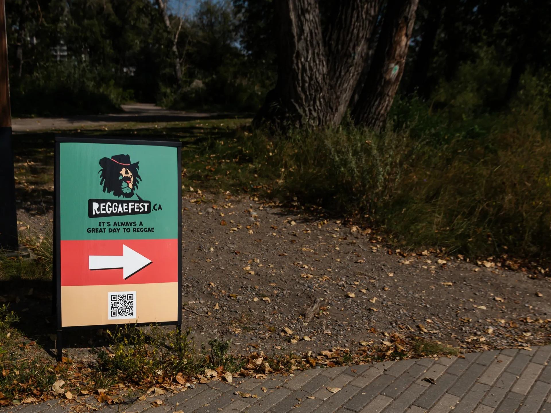 Reggaefest.ca directional sign with arrow beside dirt path and large trees.