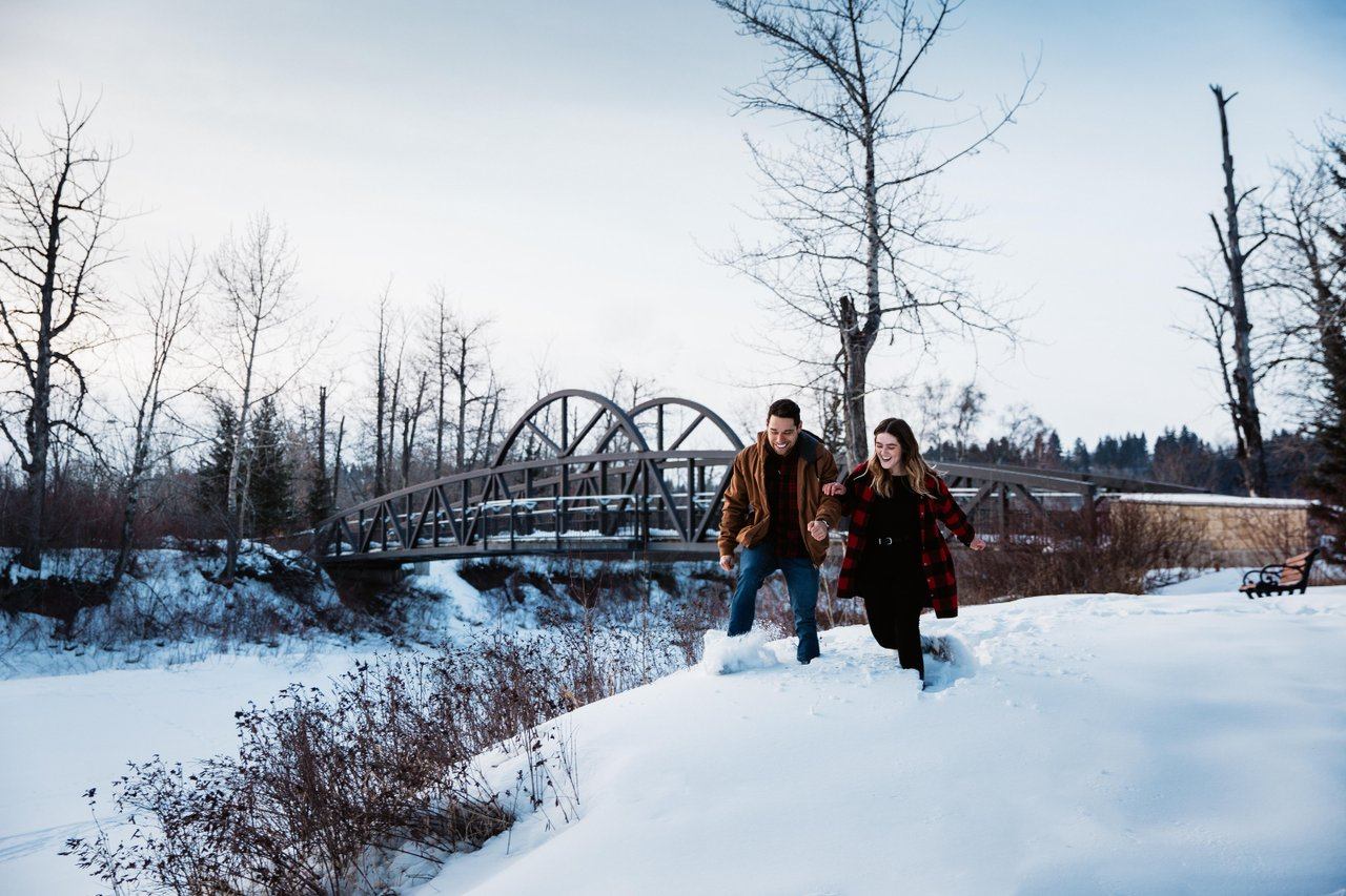 Two people walk through deep snow near a metal bridge and bare trees.