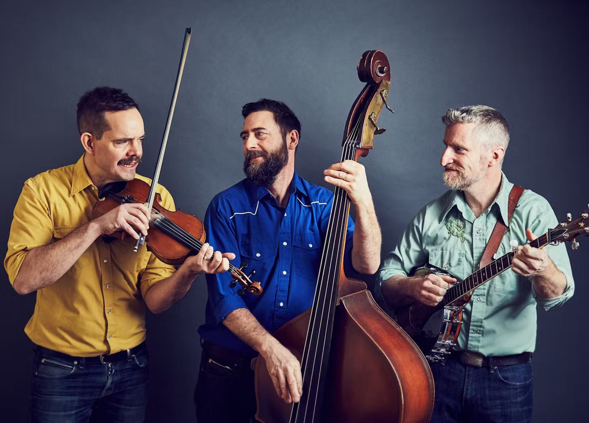Three musicians playing violin, bass, and banjo against a dark background.