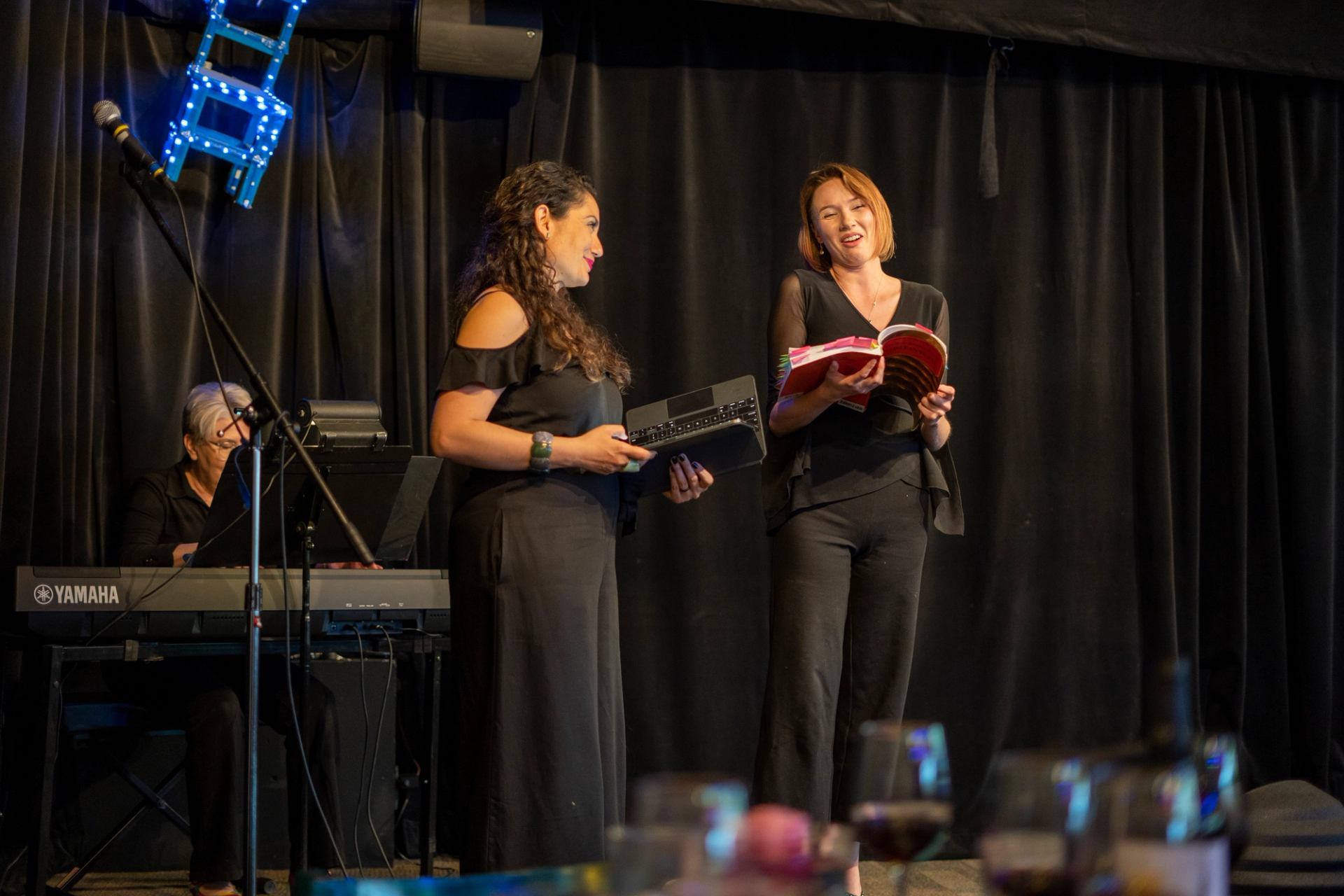Trio performs on stage with keyboard, book, and wine glasses in cozy pub setting.
