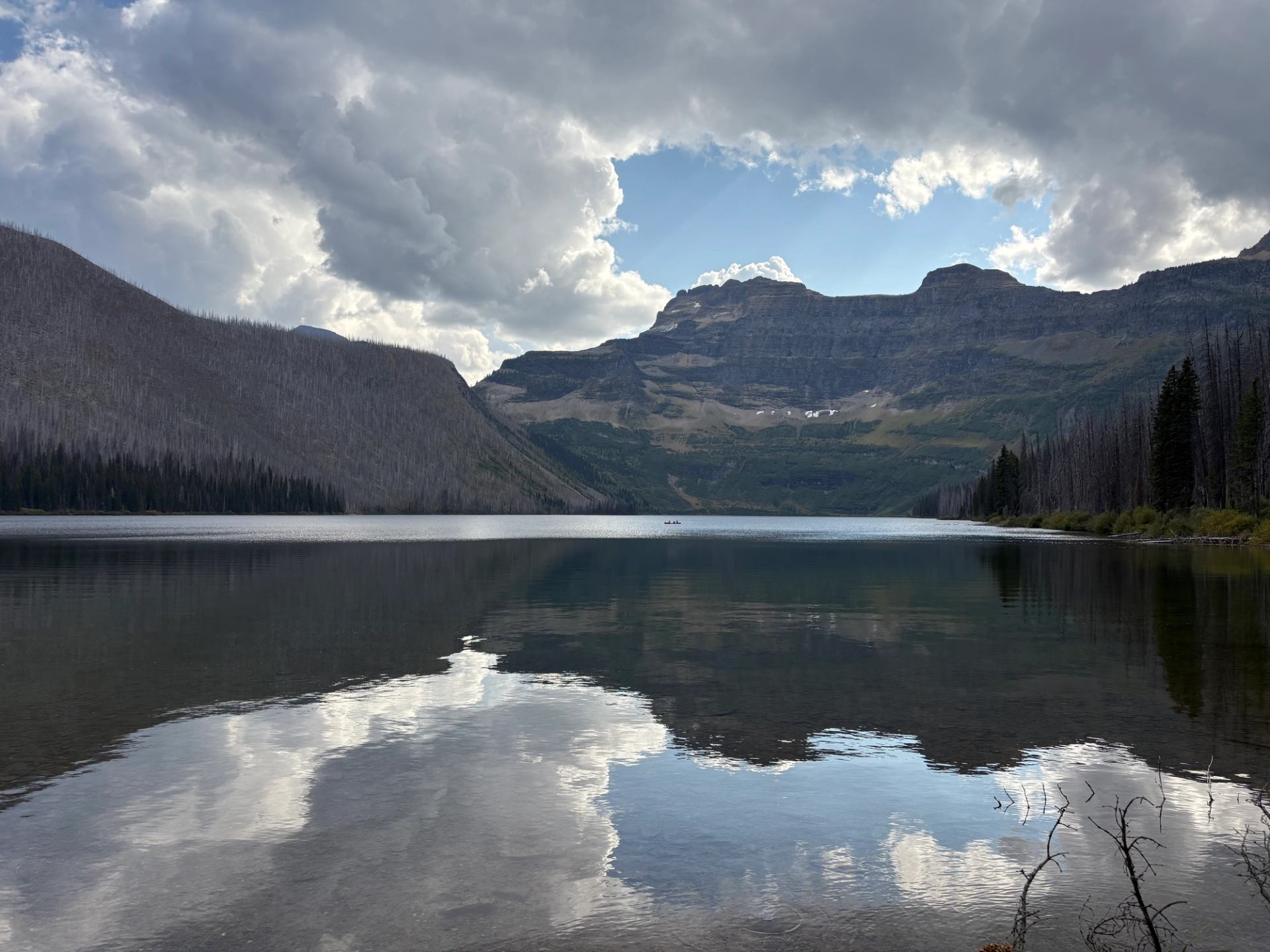 Mountain lake with steep forested slopes and clouds reflected on calm water surface.