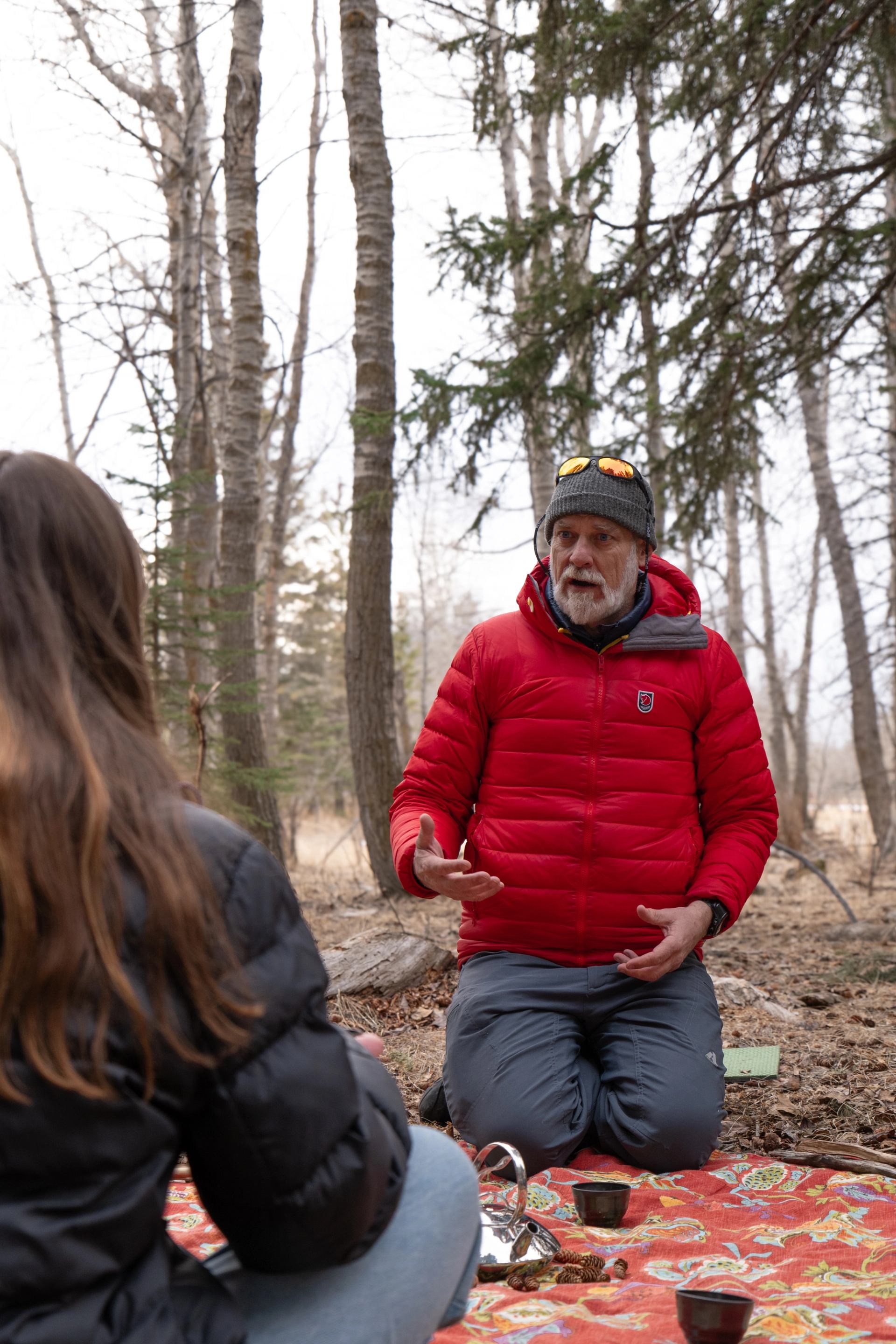 A man in a red jacket sits cross-legged on a blanket in a calm forest, talking with a woman.