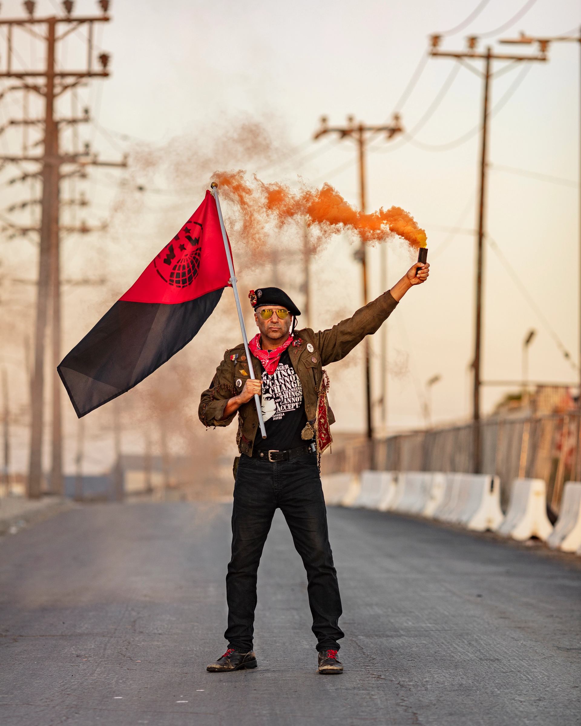 Protester stands on empty road holding red and black flag and orange smoke flare.