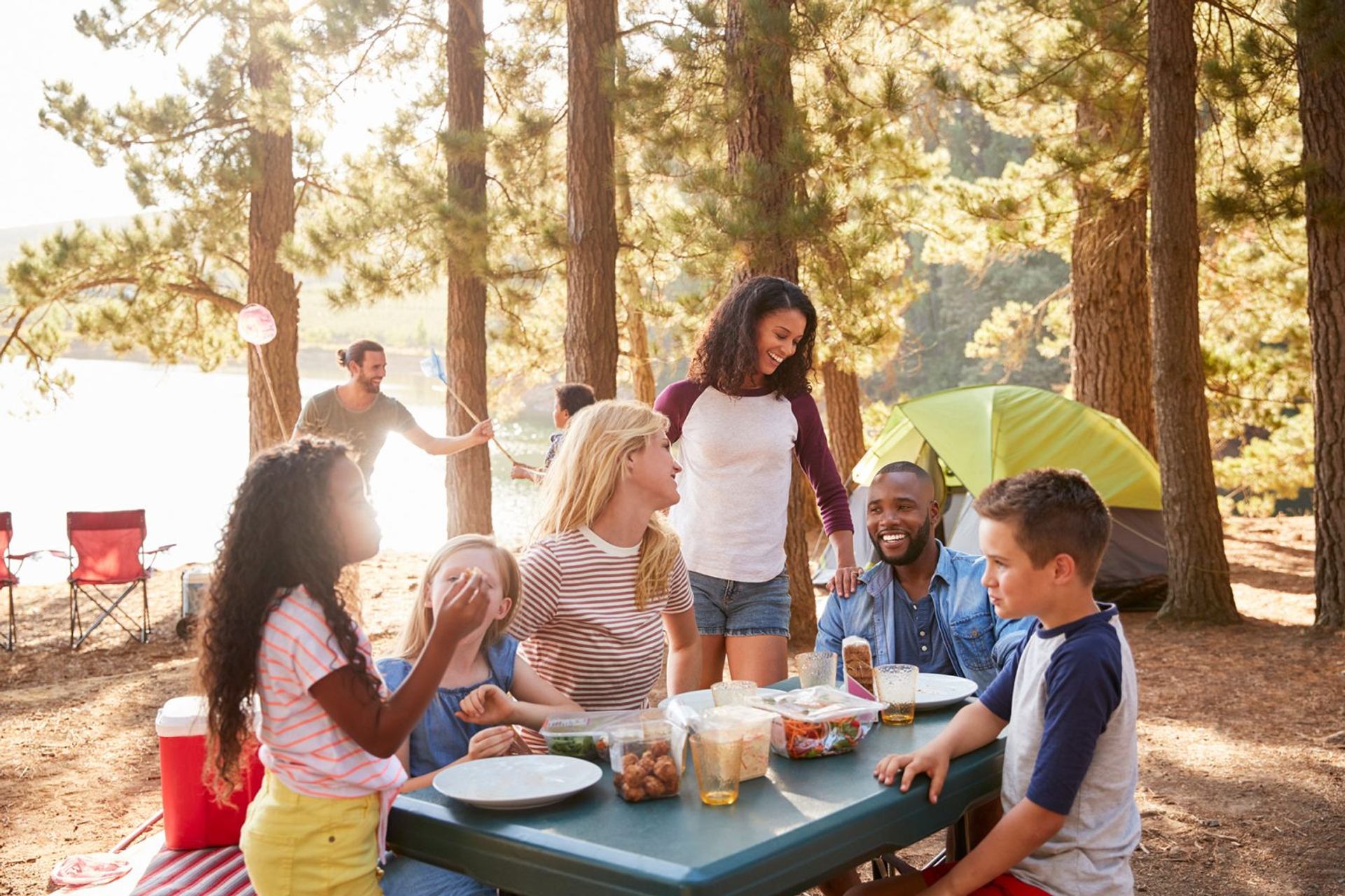 Campers gather around a picnic table at a forested campsite near tents and a lakeside setting.