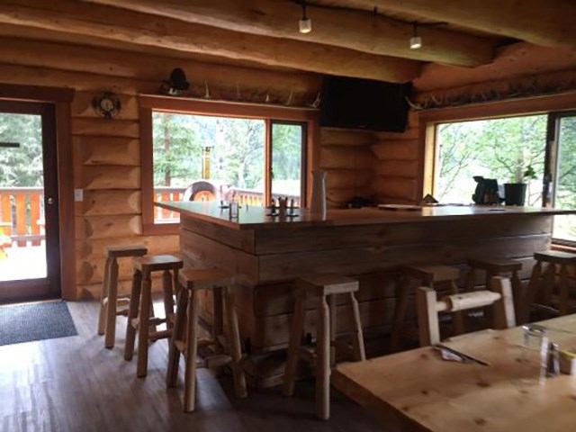 Cozy log cabin interior with a wooden bar, stools, and large windows overlooking nature.