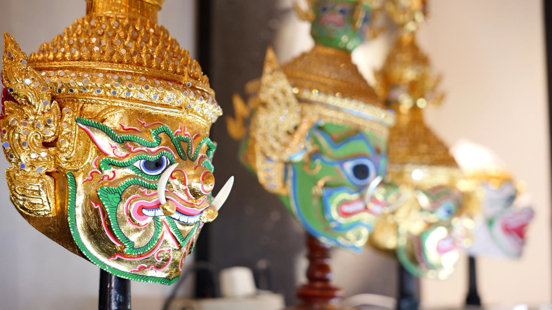 Ornate golden Thai Khon masks with colourful painted faces displayed on stands indoors.
