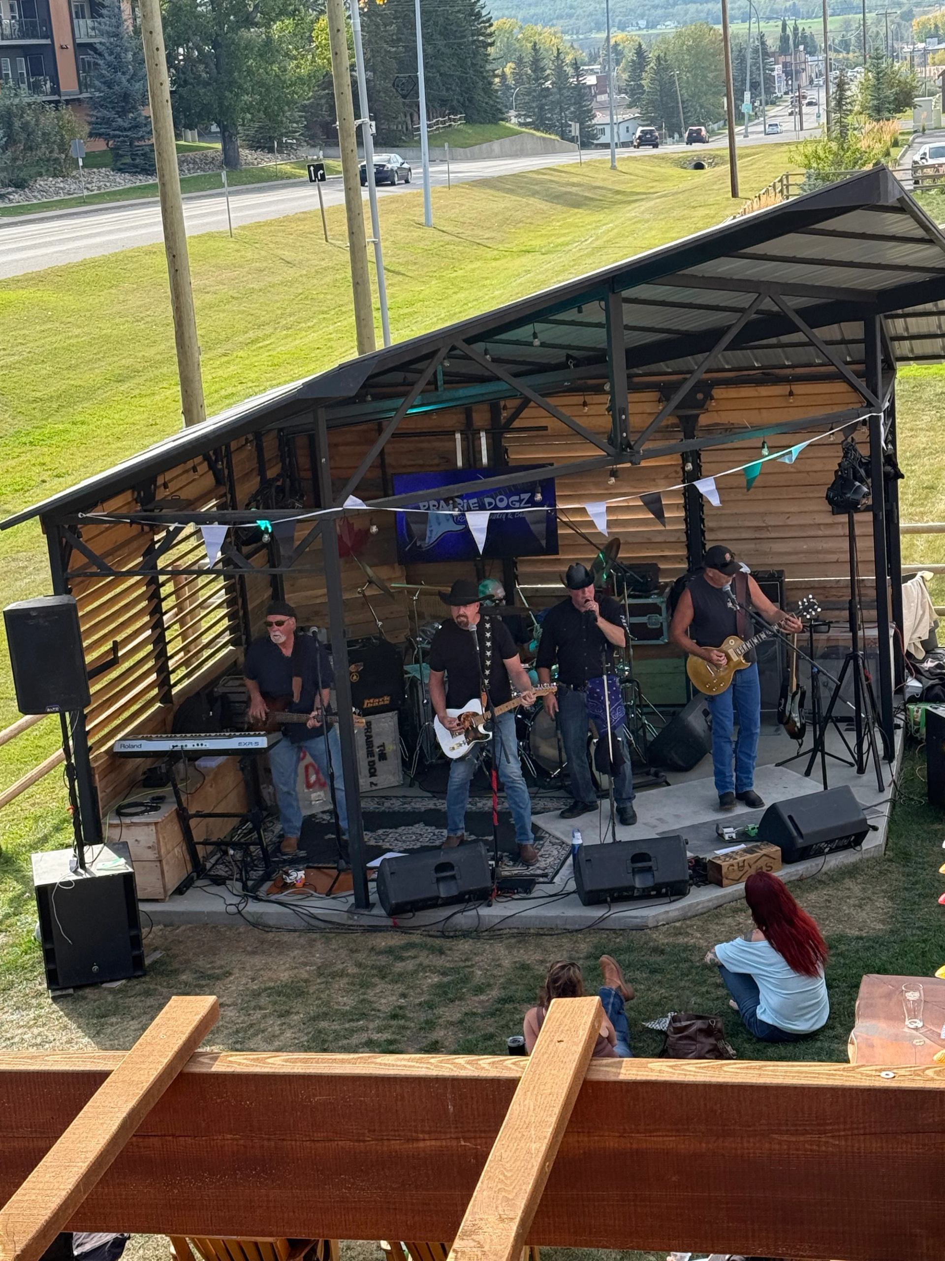 The band Prairie Dogz performs on a covered outdoor stage with an audience on the grass, a road and hills in the background.