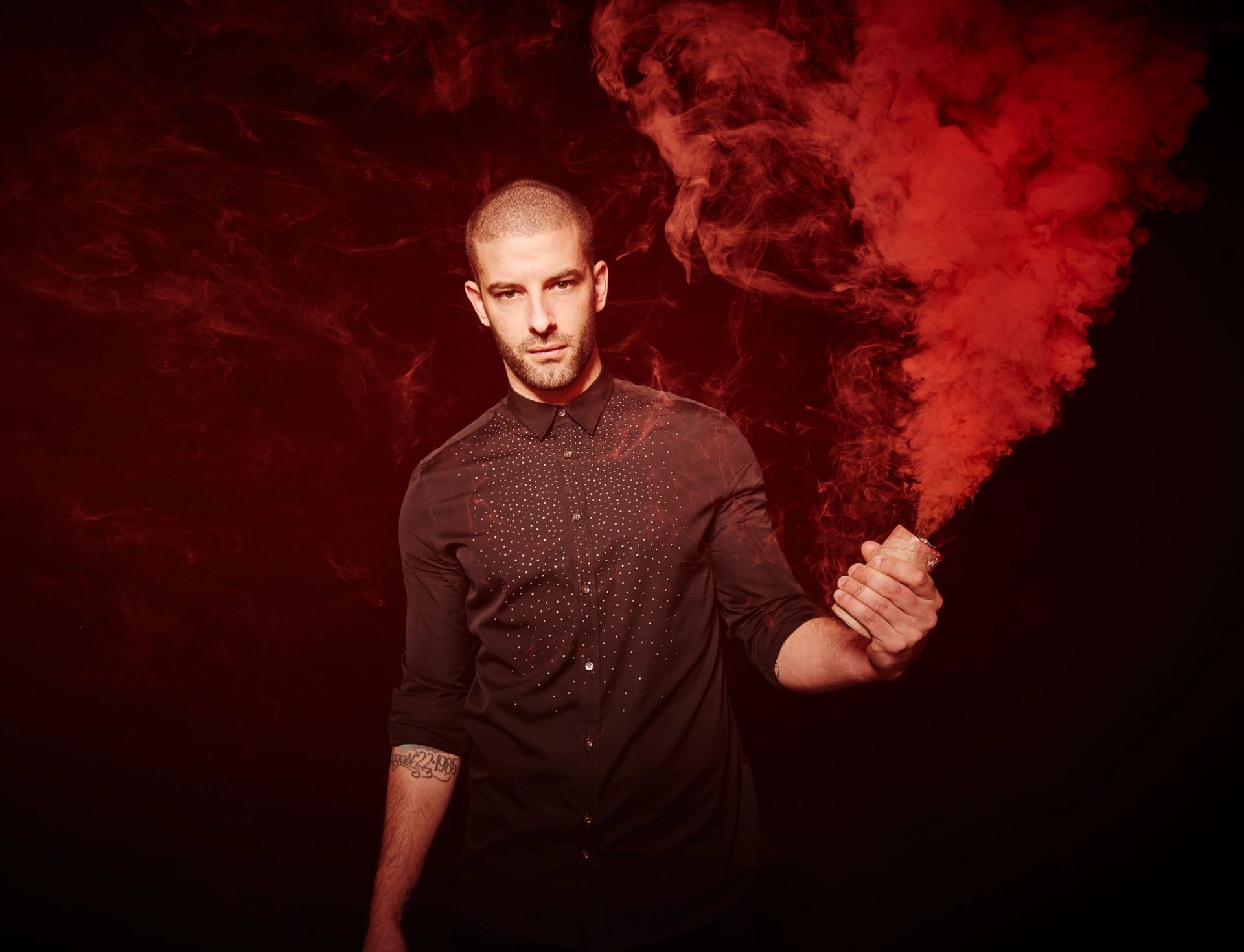 Person in dark shirt holds red smoke flare against dark red smoky background.