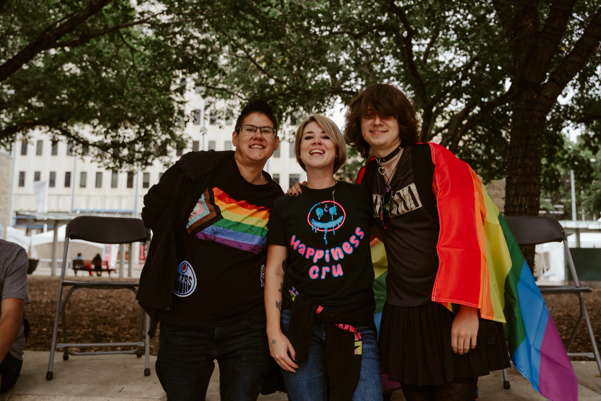 Three people pose outdoors under trees, one draped in a rainbow flag.