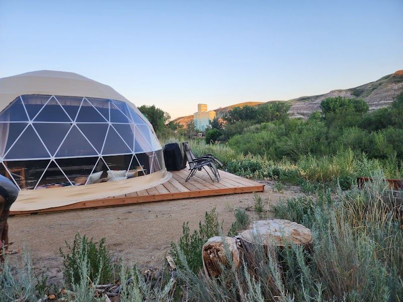 Geodesic dome tent on wooden deck with lounge chairs in grassy valley landscape.