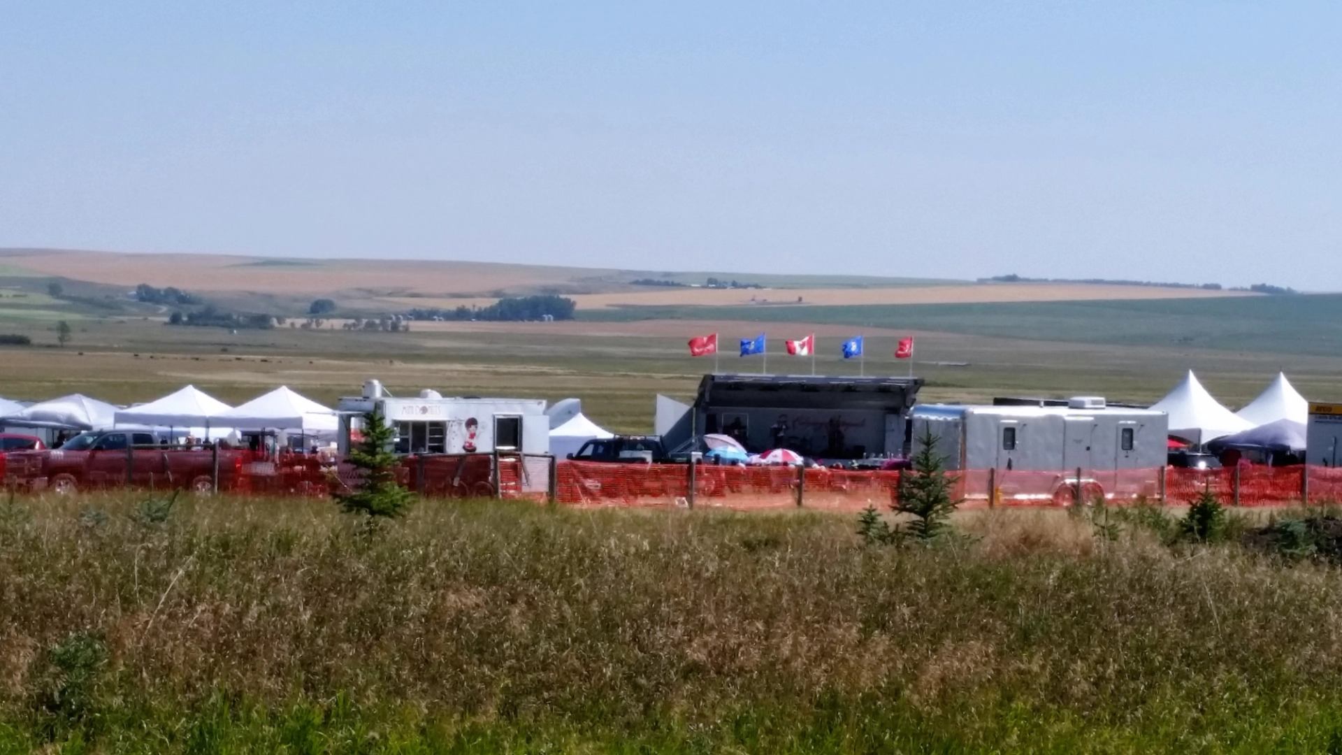 Festival tents and parked vehicles with Canadian flags on poles in rural fields.