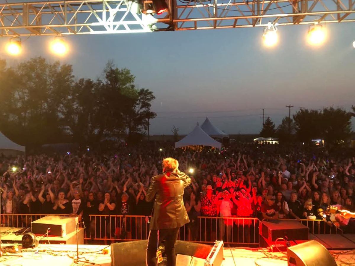 Singer on outdoor stage performing for large crowd at dusk under bright lights.