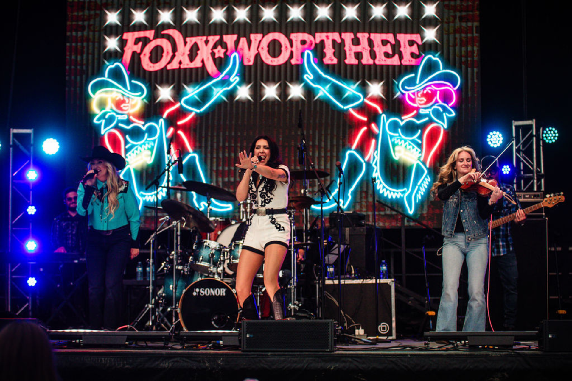 Foxx Worthee on stage with neon lights behind. Two singers and a fiddle player with a backing band.